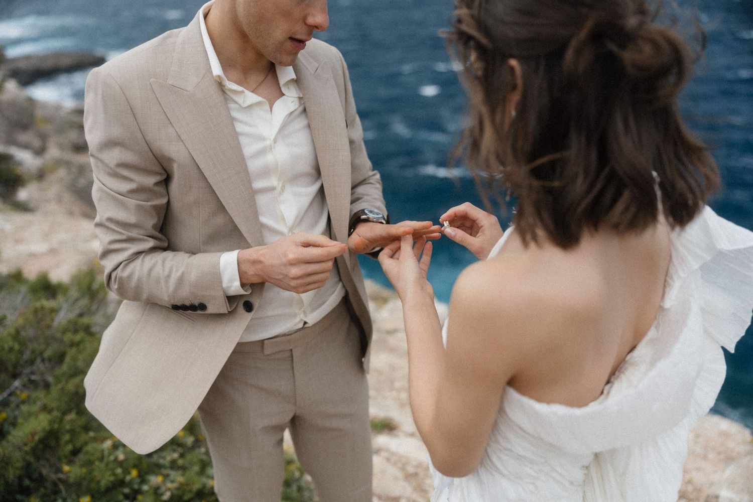 Couple exchanging wedding rings on a cliffside overlooking the ocean, groom in beige suit, bride in white dress.
