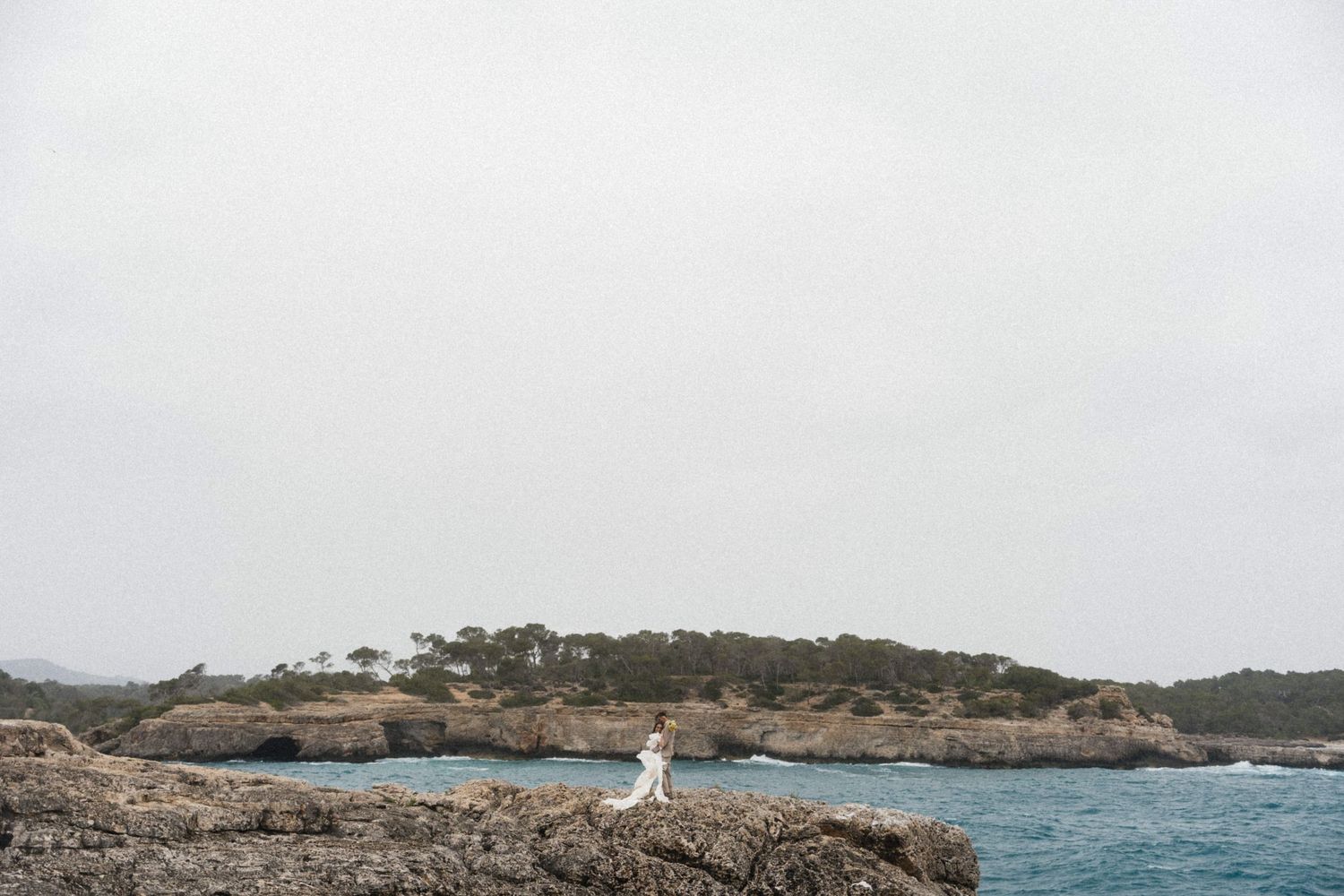 Bride in white dress standing on rocky coastal cliffs with turquoise ocean and tree-covered island backdrop.