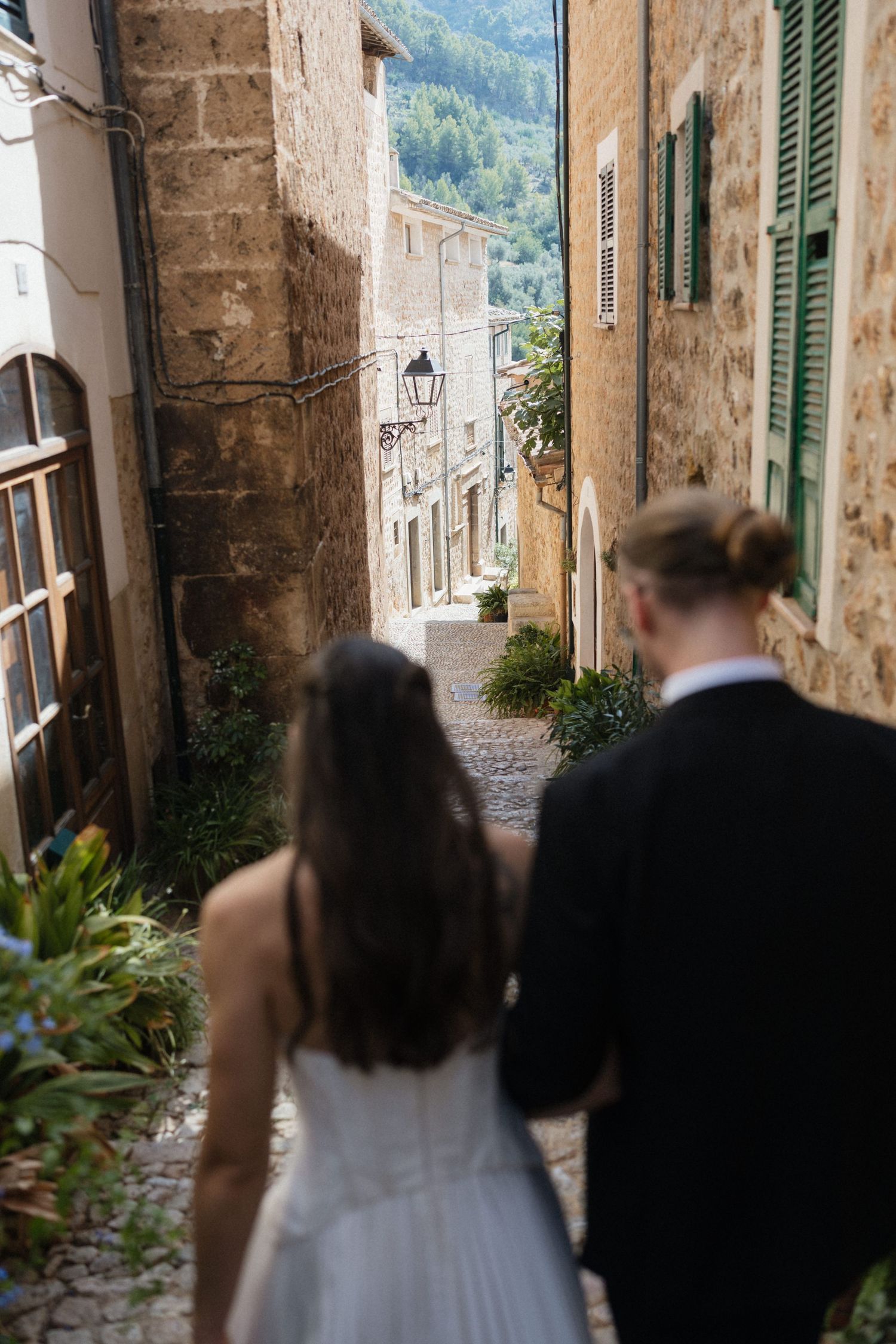 Bride in a white dress and groom in a dark suit walk away down a narrow stone alley lined with greenery in a charming Mediterranean village.