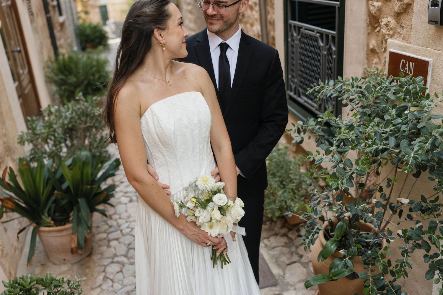 Bride in a strapless white wedding dress holding a white bouquet embraces a groom in a dark suit on a cobblestone path among potted plants.