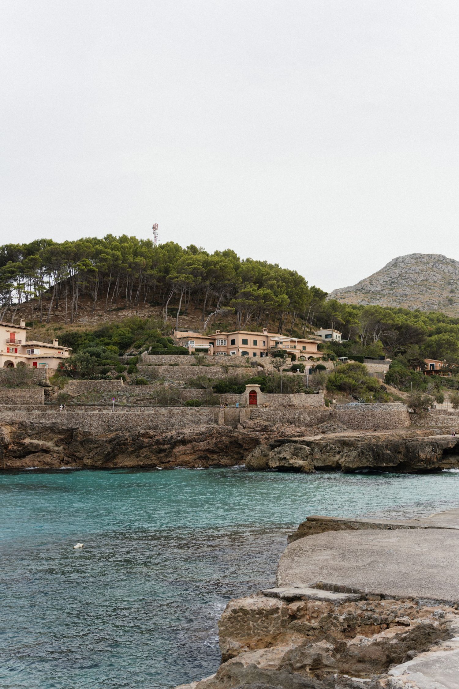 Turquoise Mediterranean cove with rocky shoreline and stone-walled villas nestled among pine trees on a hillside beneath a rugged mountain.