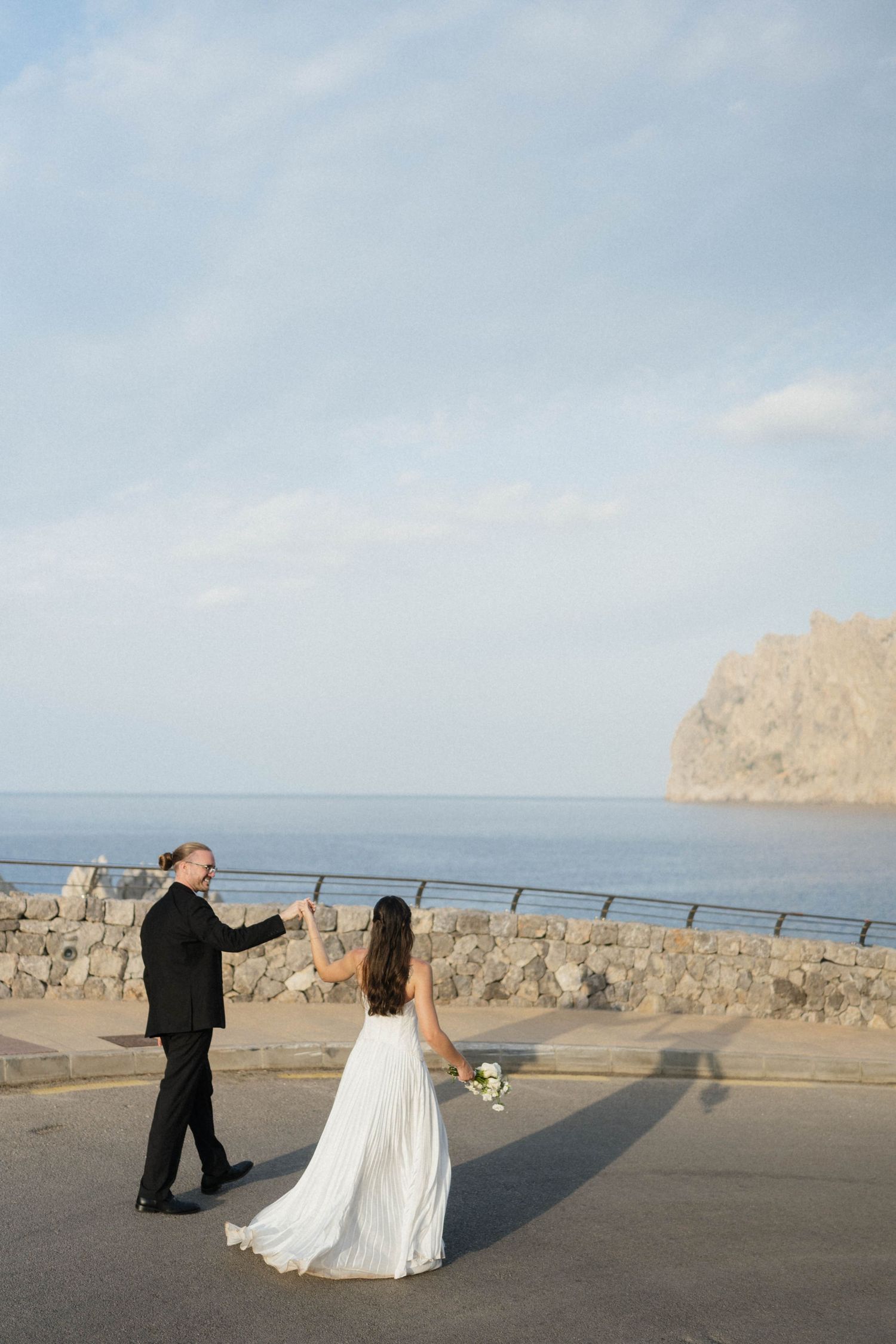 Bride in a white gown and groom in a black suit dance hand in hand on a seaside road with a rocky headland behind.