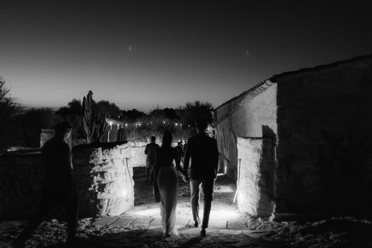 A black and white night photo of three people walking along a narrow stone path between rustic walls under a starry sky.