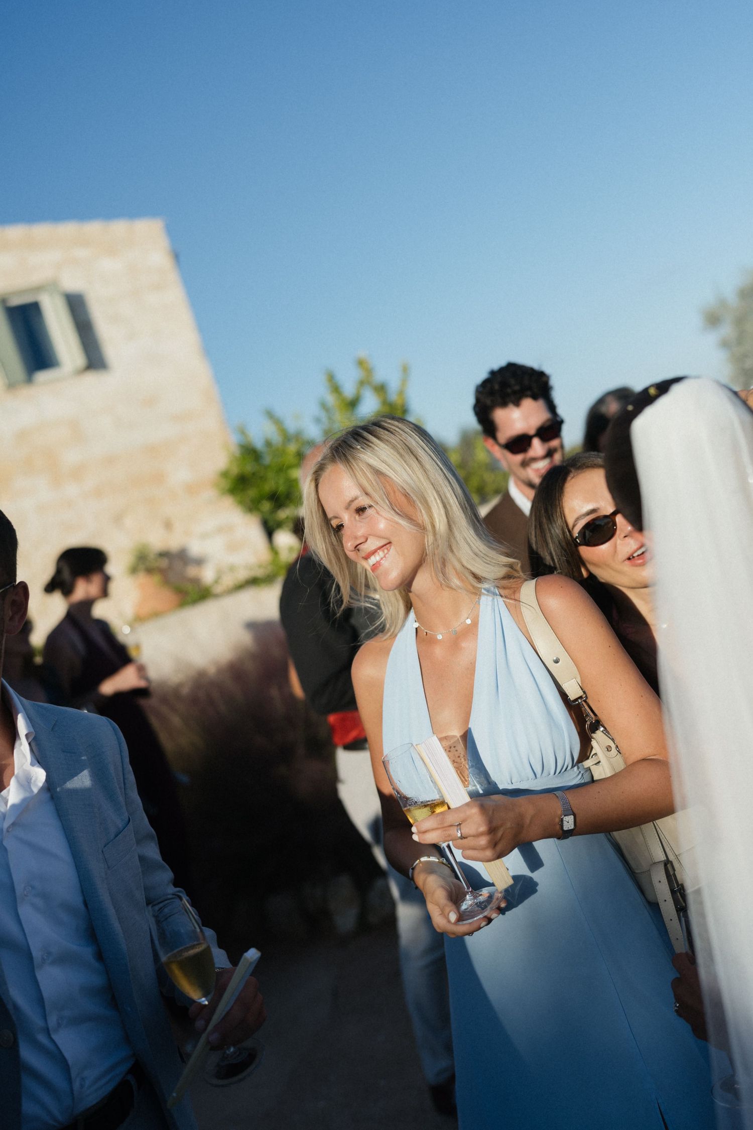 Smiling woman in a light blue dress holds a champagne glass among wedding guests at a sunny outdoor reception.