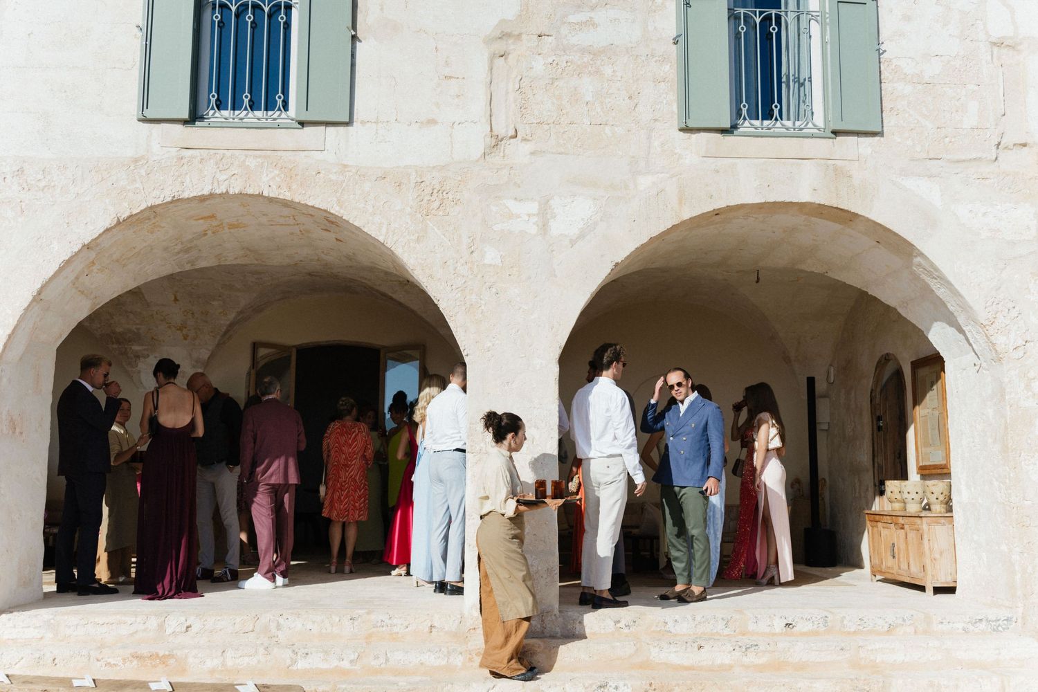 Wedding guests in elegant attire gather beneath the stone archways of a rustic Mediterranean building with green shutters.