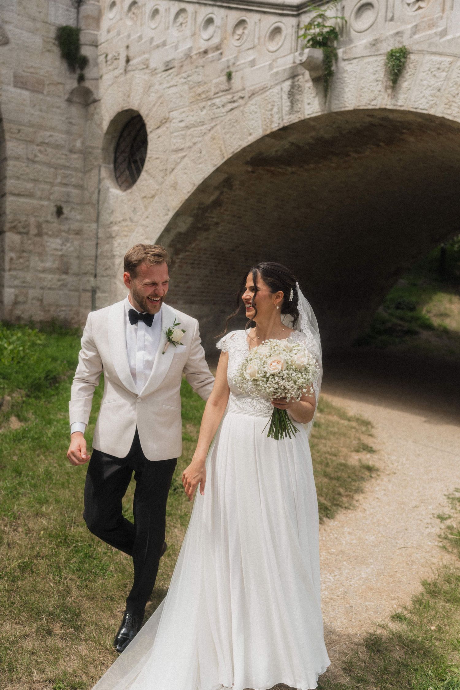 Smiling bride and groom walking hand in hand near stone arch bridge outdoors.