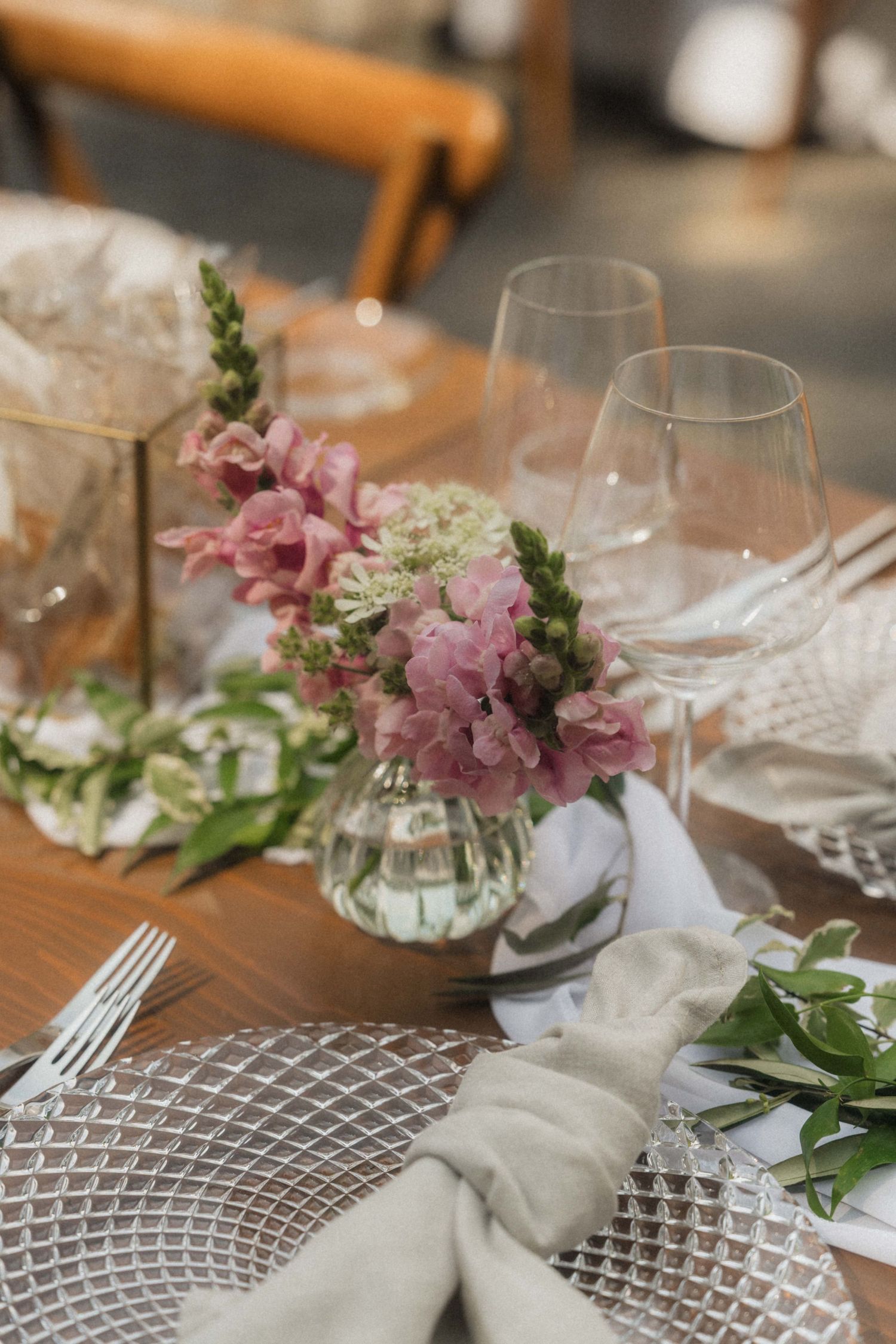 Elegant wedding table setting with pink snapdragon flowers, crystal glassware, and linen napkins on wood table.