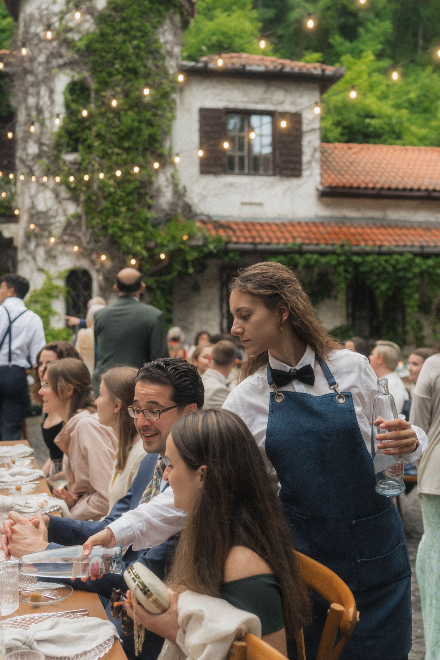 Waitress serving drinks to wedding guests at an outdoor reception with string lights and greenery.
