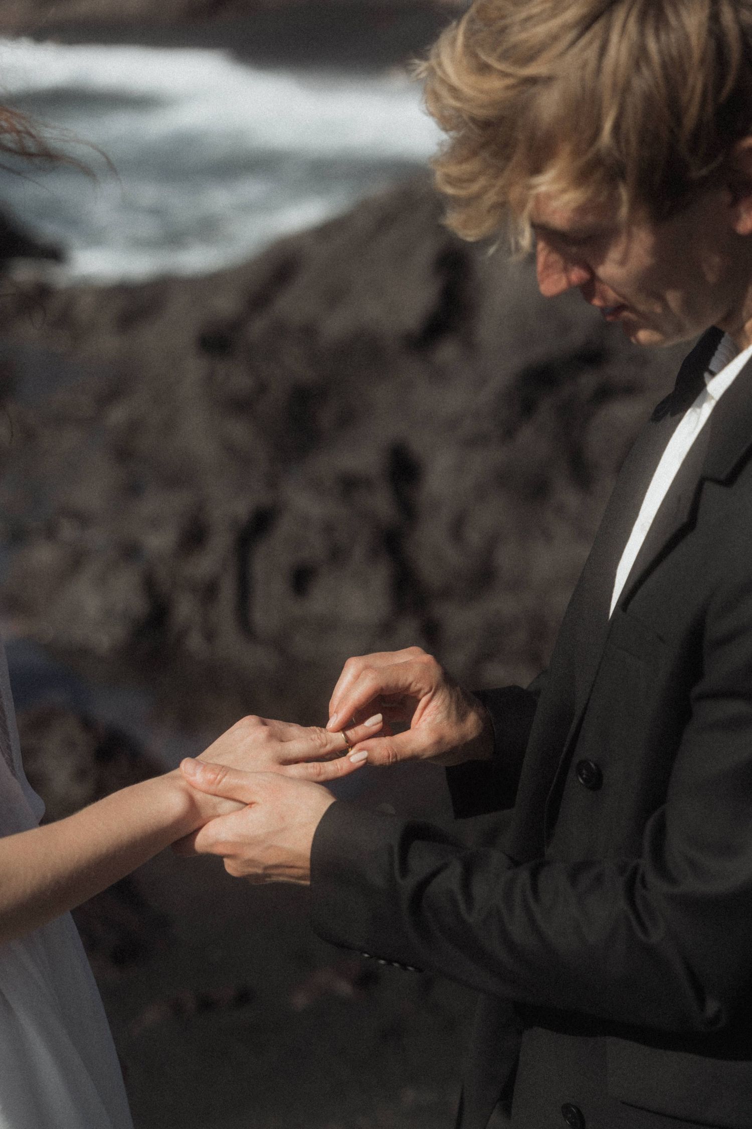 Groom placing wedding ring on brides finger during intimate outdoor ceremony near rocky coastline.