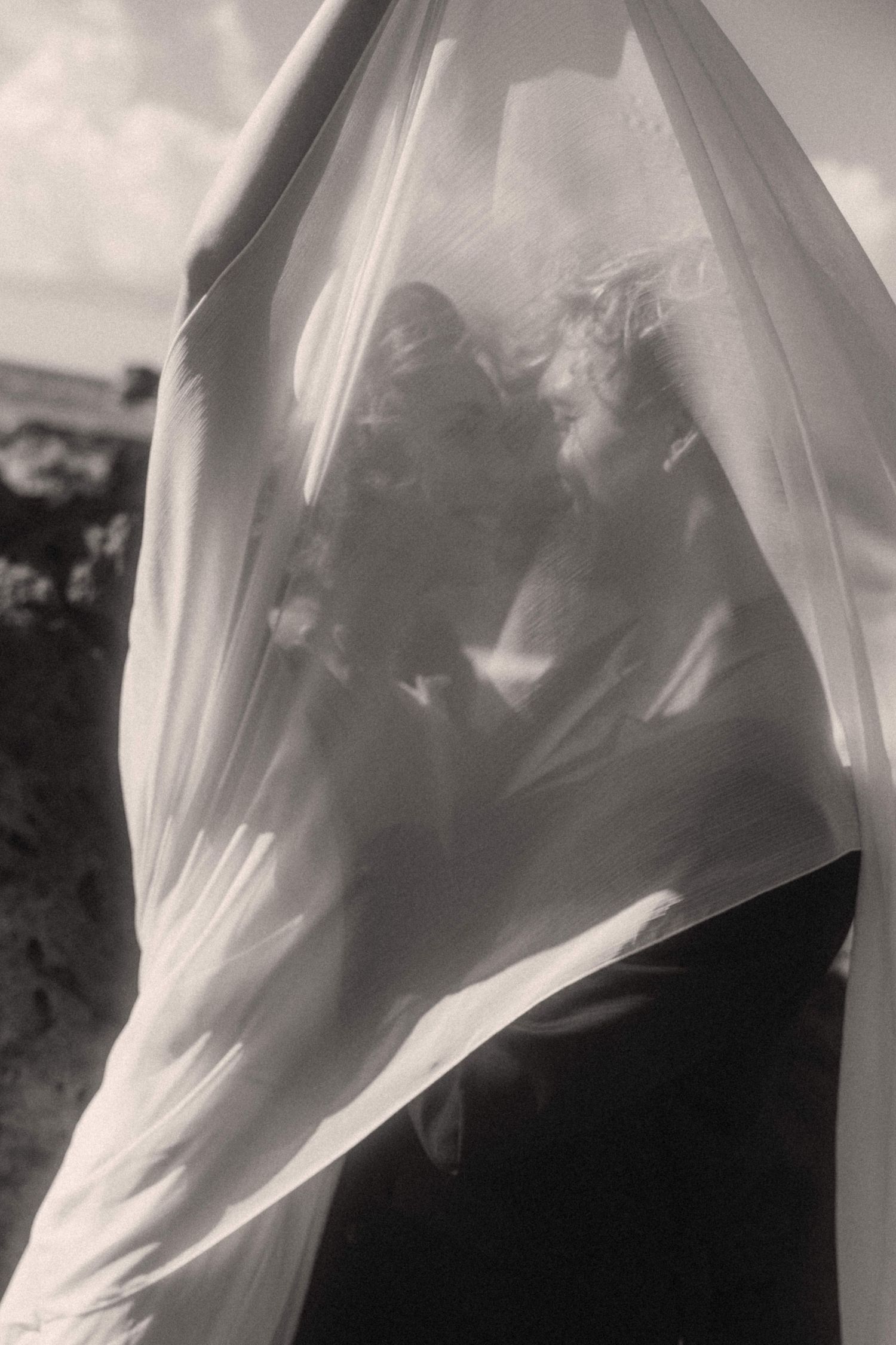 Romantic black and white photo of couple kissing beneath a sheer flowing veil outdoors.