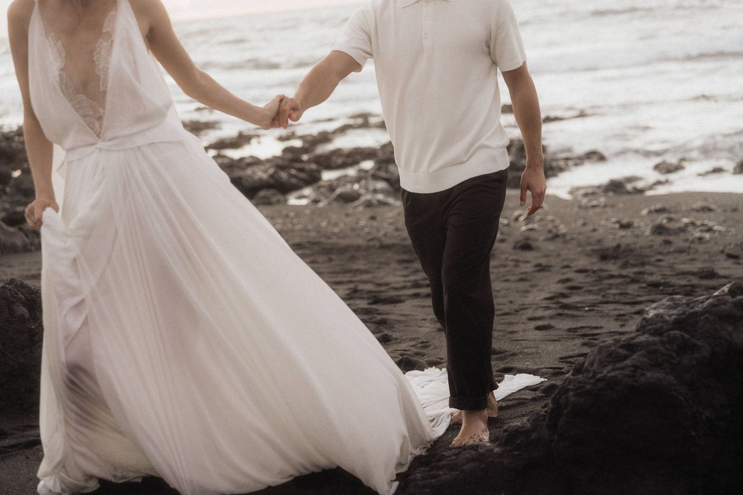 Bride in flowing white lace gown holds groom's hand walking on rocky beach with ocean waves behind them.