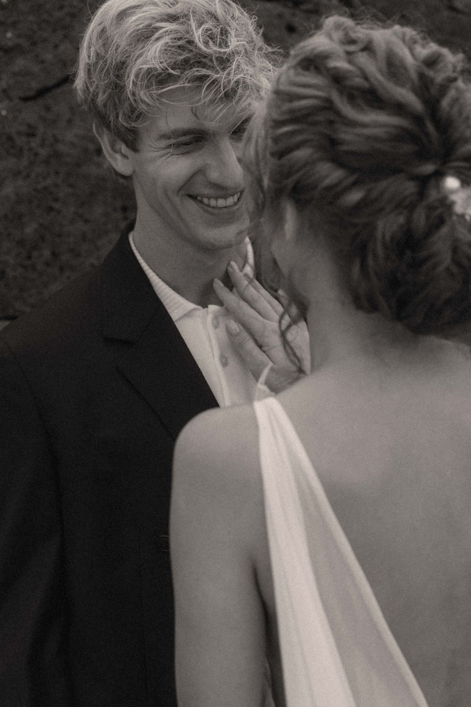 Groom smiling joyfully at bride in black and white wedding photo, bride in backless white gown touching his face.