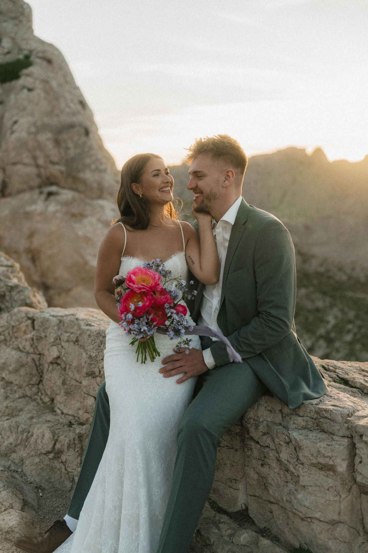 A smiling bride and groom pose on rocky mountain terrain at golden hour, bride holding a vibrant pink bouquet.