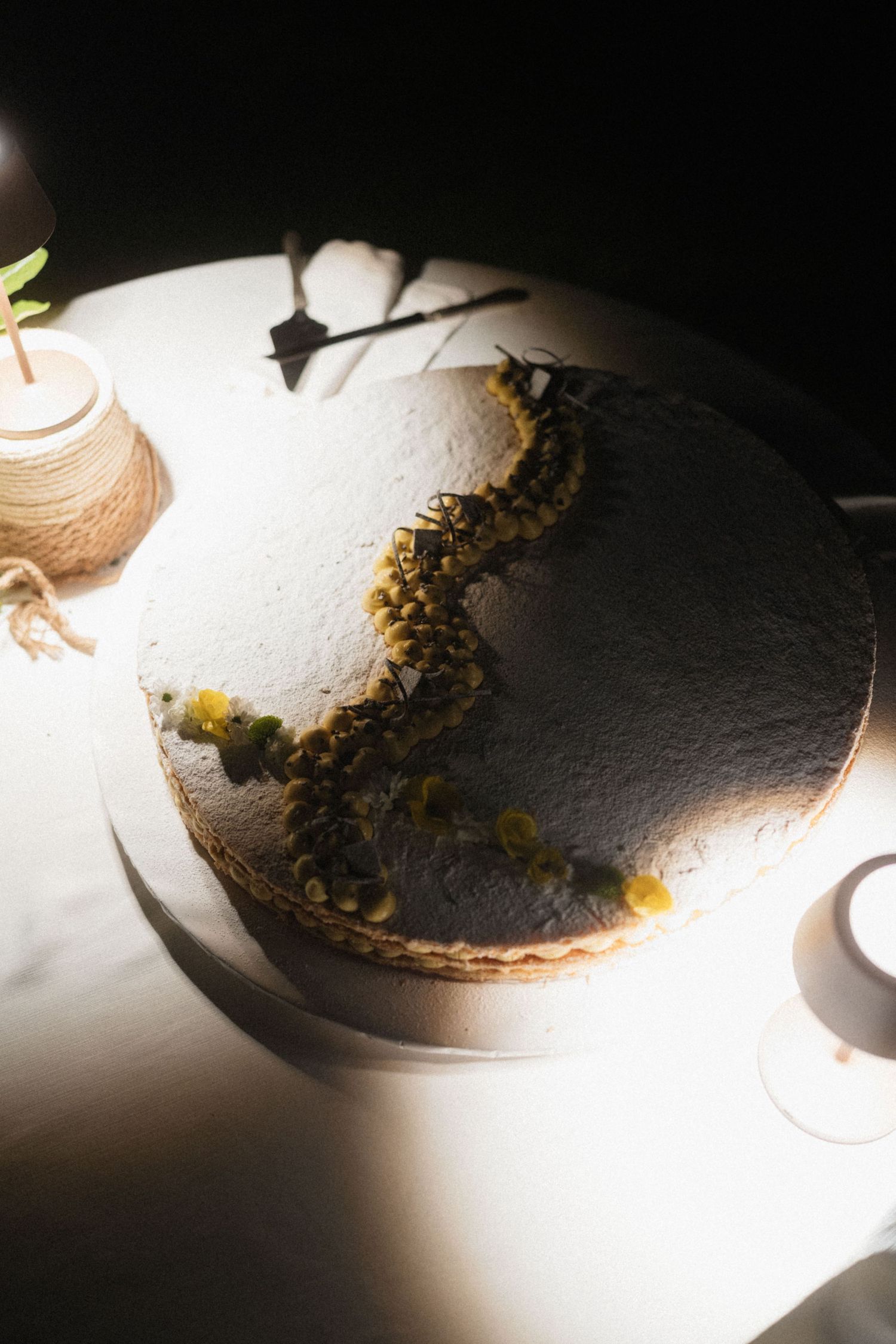 Artistic wedding cake with dark chocolate decoration and floral accents illuminated by dramatic spotlight.