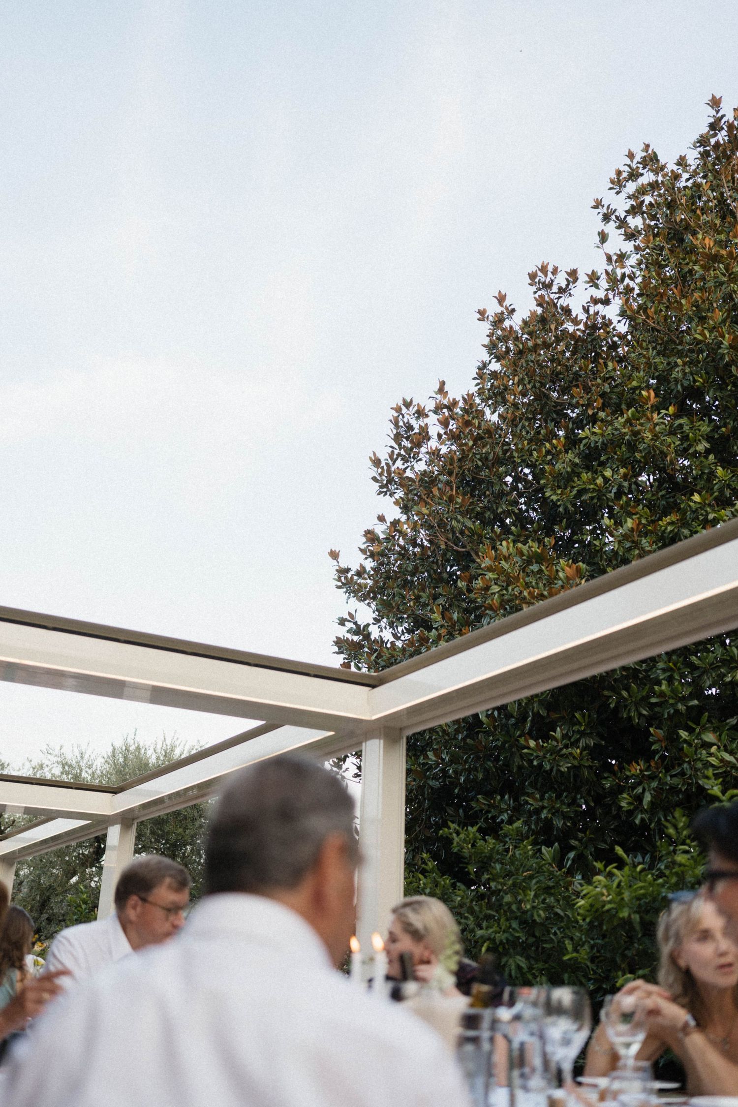 Guests dining outdoors under a white pergola surrounded by lush green trees at an elegant garden wedding reception.