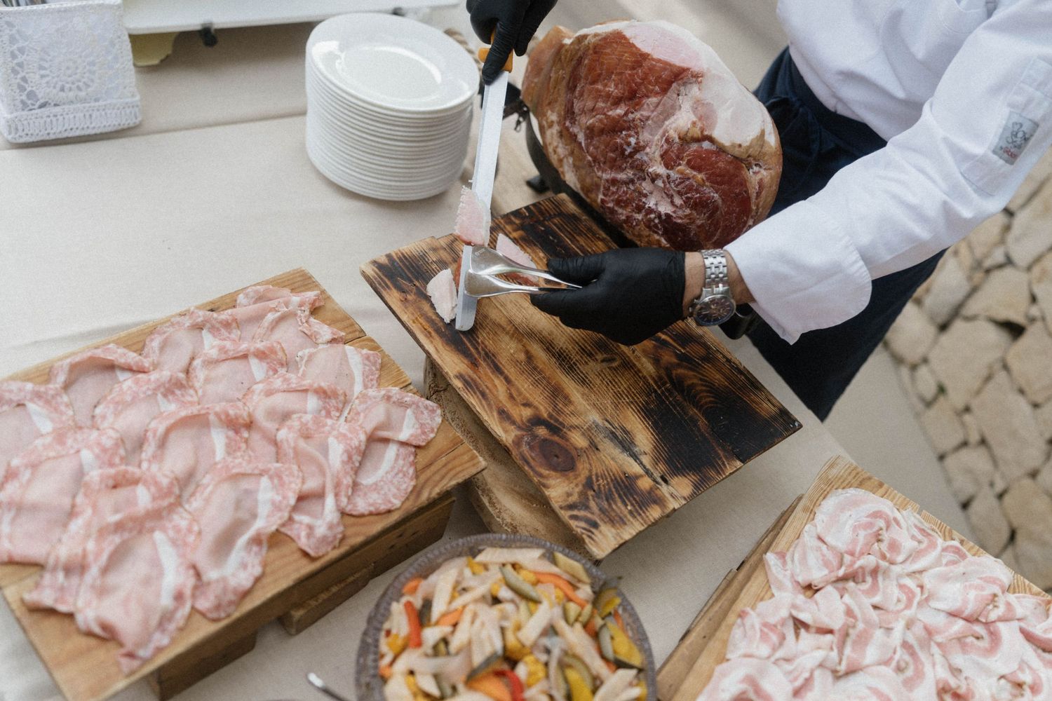 Chef in black gloves slicing cured ham on wooden board with arranged charcuterie and pickled vegetables nearby.