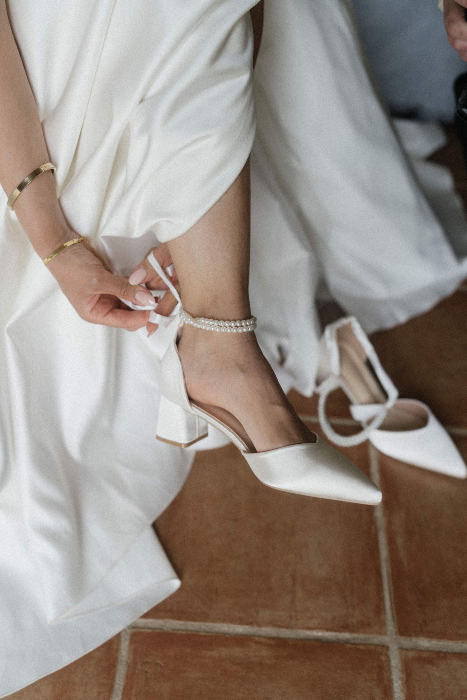 Bride fastening pearl ankle strap on white block heel wedding shoes while seated.
