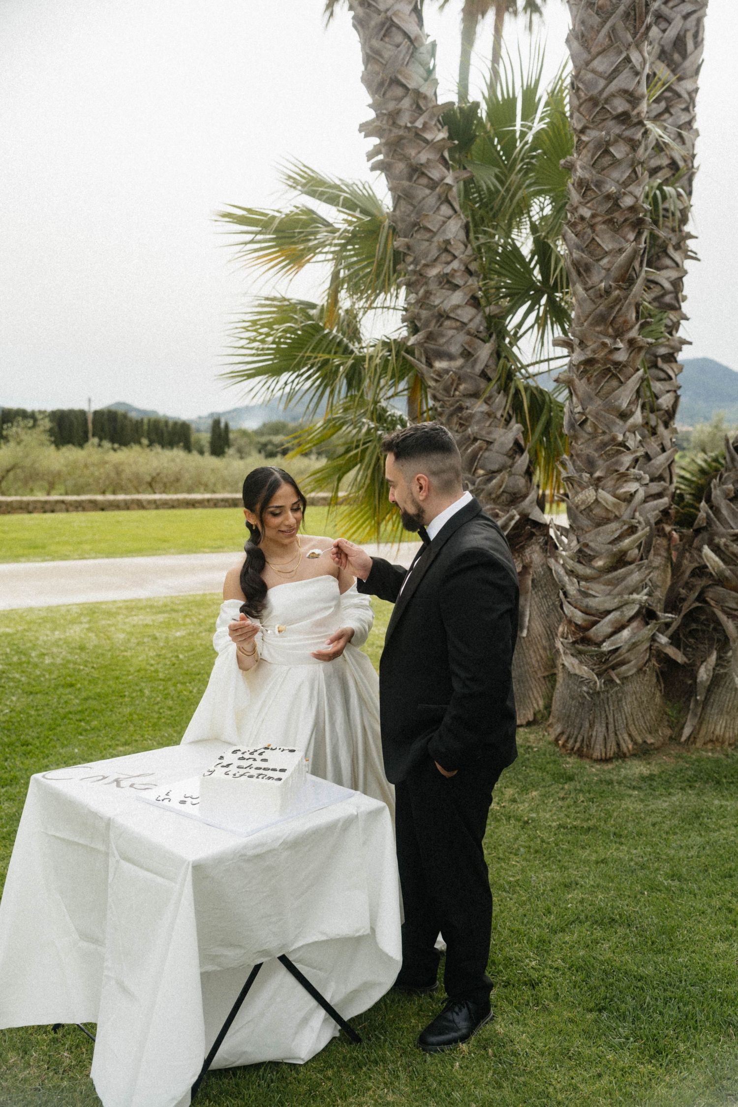 Bride and groom exchanging rings at outdoor wedding ceremony table with palm trees backdrop.