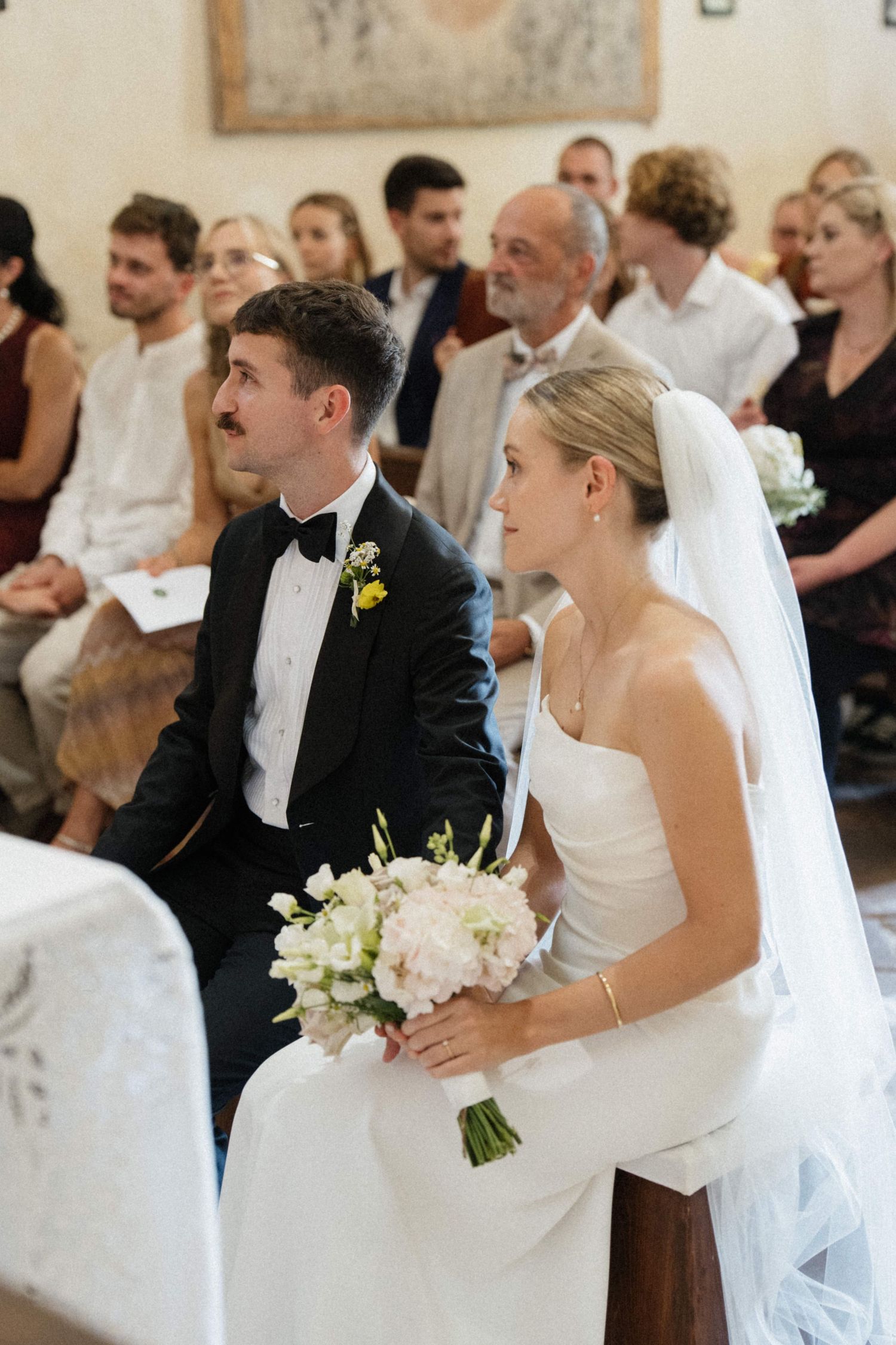 Bride and groom sit together during church wedding ceremony, bride holding white bouquet, guests seated in pews behind them.