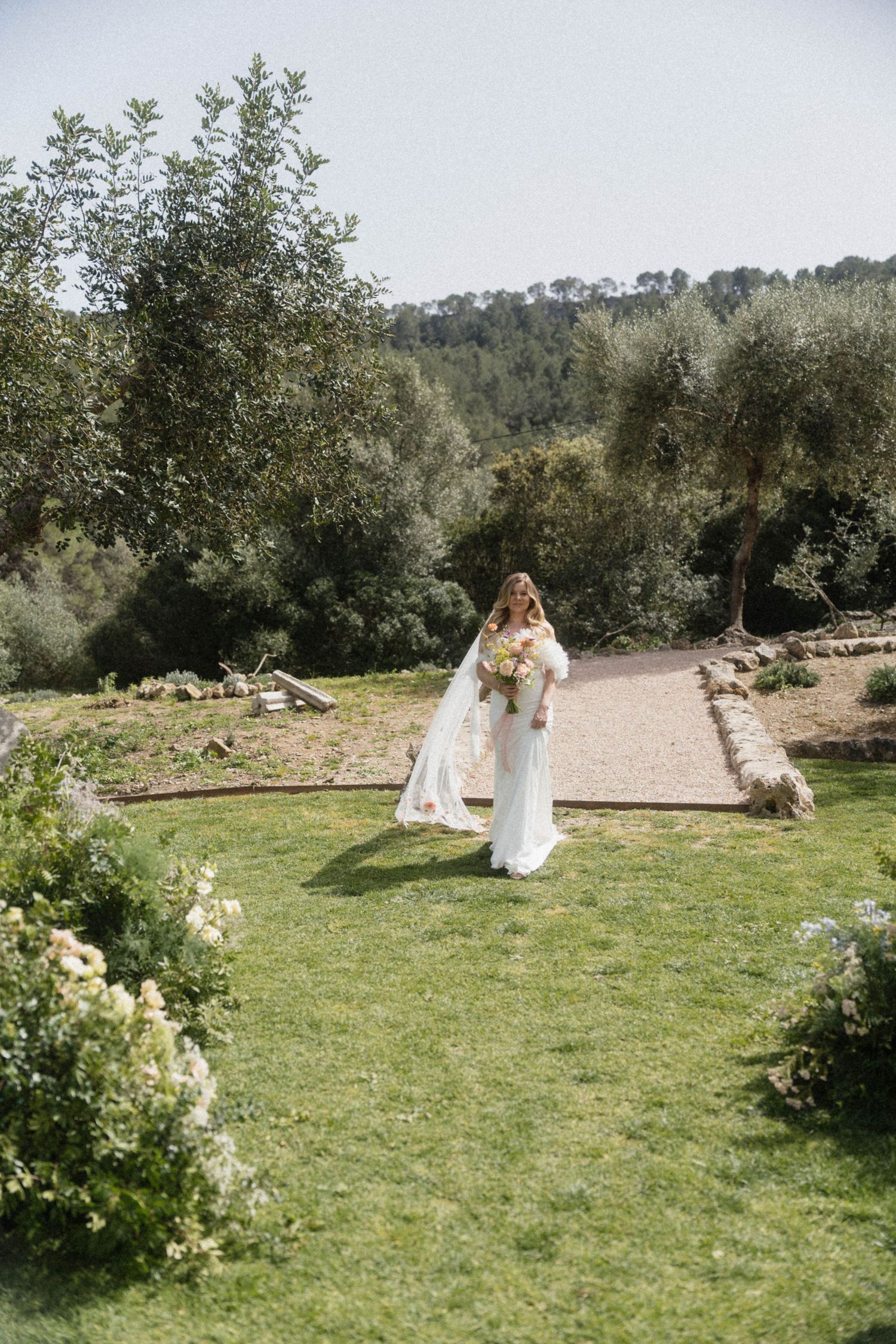 Bride in a fitted white wedding gown with feathered cape carrying a bouquet in a lush garden setting.