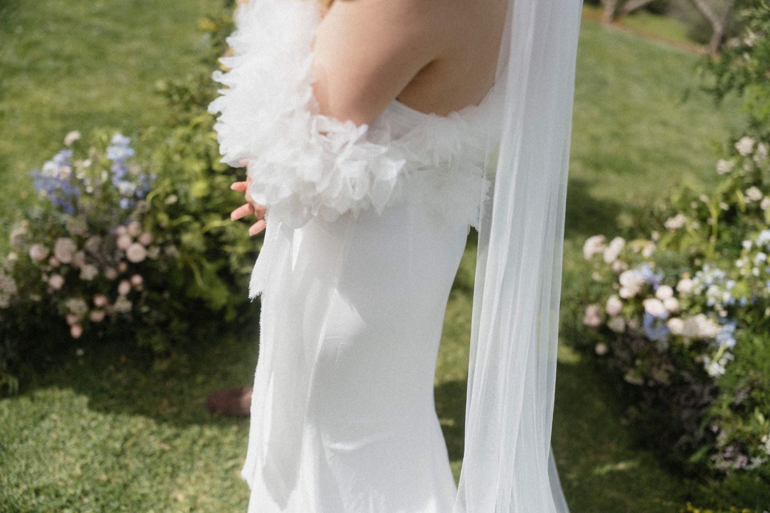 Close-up of a bride in a white off-shoulder wedding gown with ruffled sleeves and flowing veil outdoors.