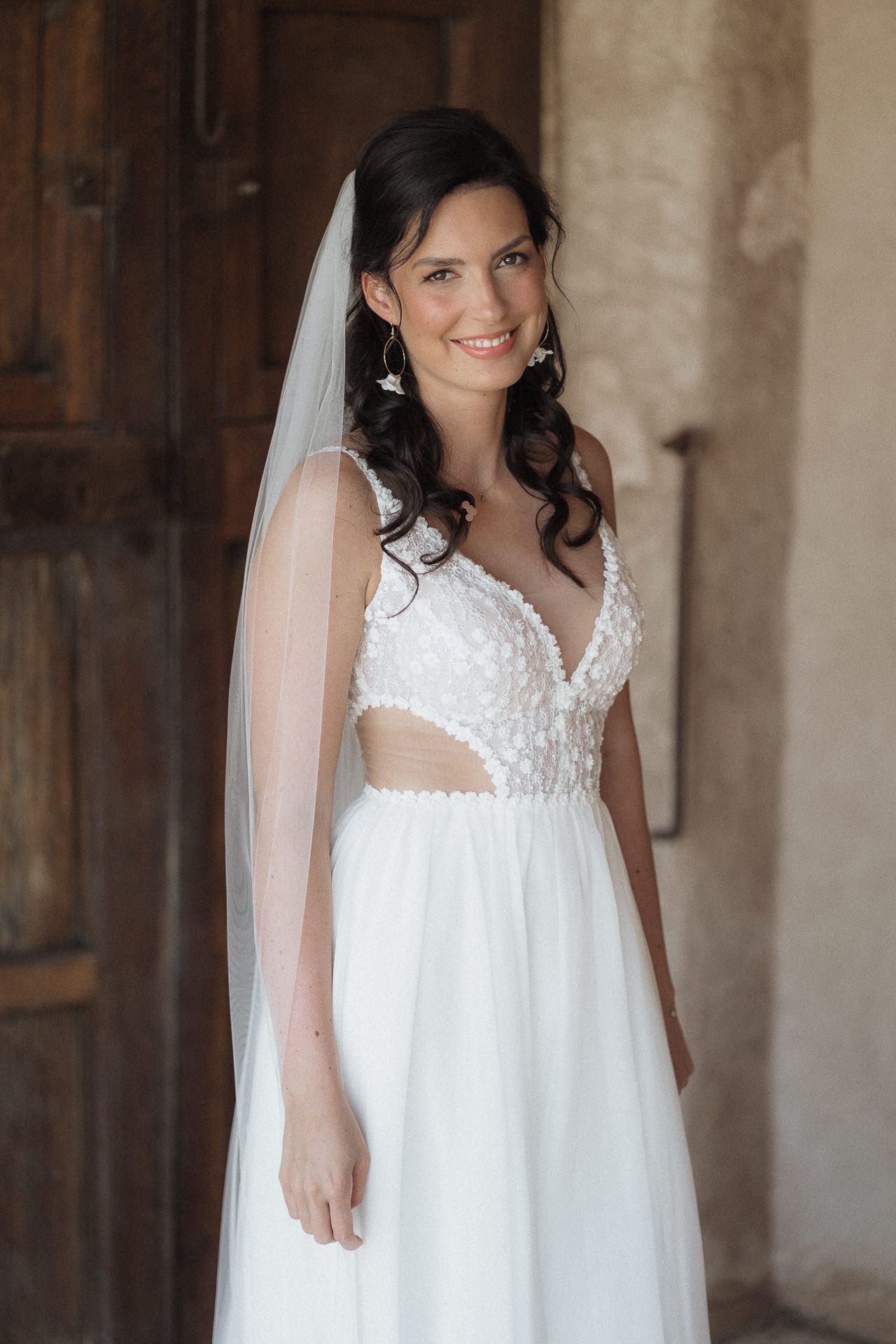 Smiling bride in a white floral appliqué wedding gown with cutouts and veil, posing near a rustic wooden door.
