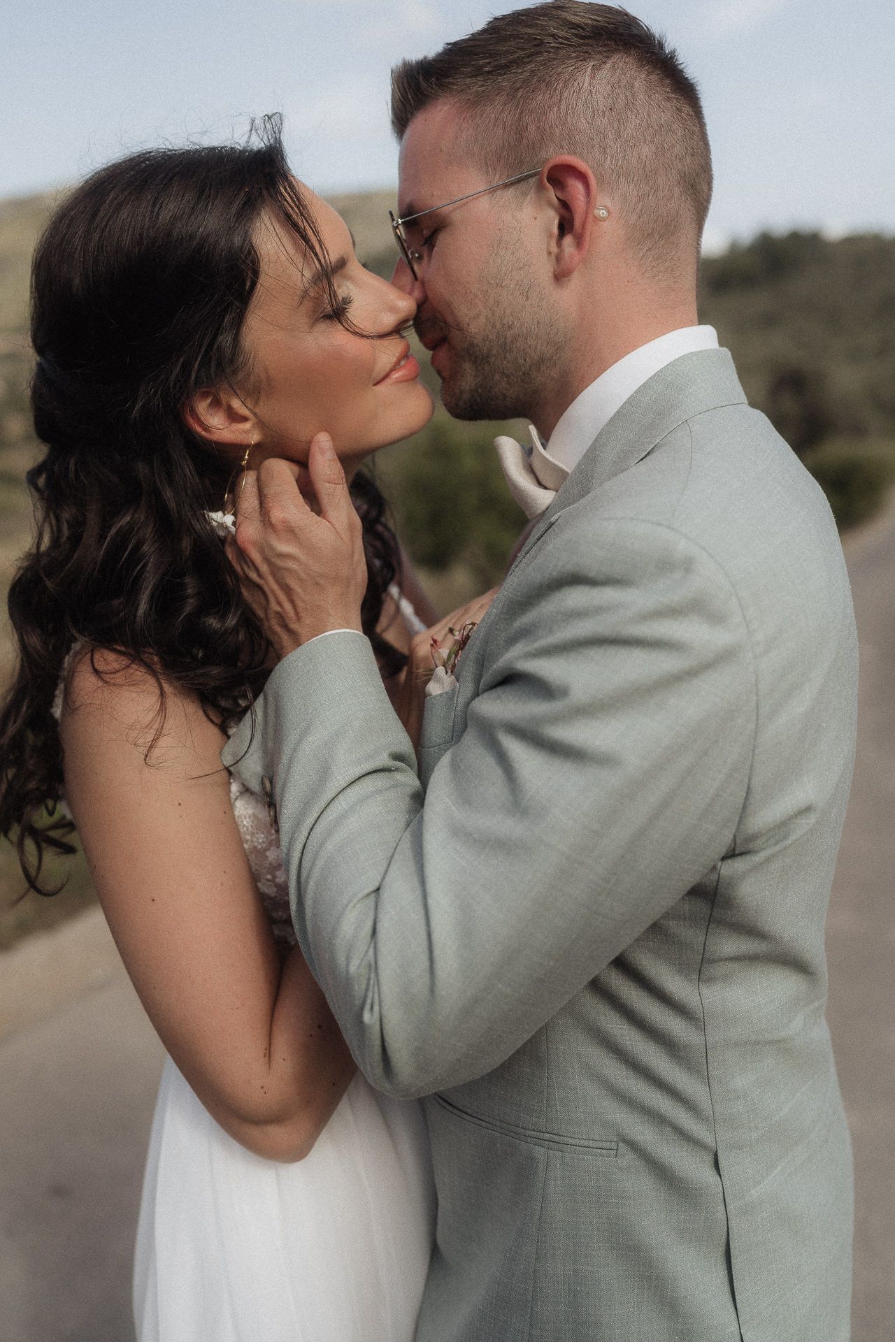 A bride and groom share a romantic almost-kiss moment outdoors, dressed in white gown and light gray suit.