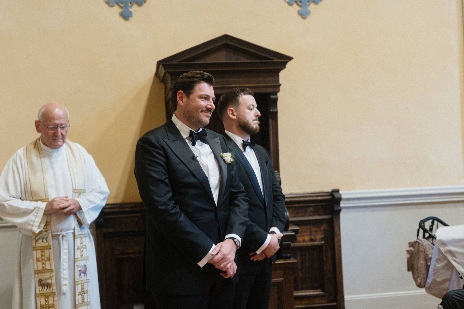 Two grooms in black tuxedos smile at altar with priest during wedding ceremony.