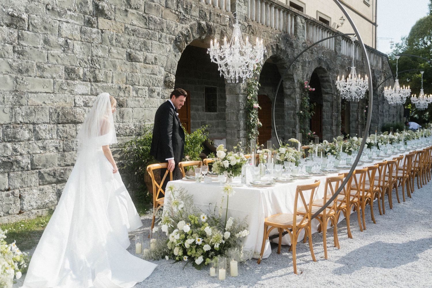 Bride and groom admiring long outdoor wedding reception table adorned with white flowers and crystal chandeliers.