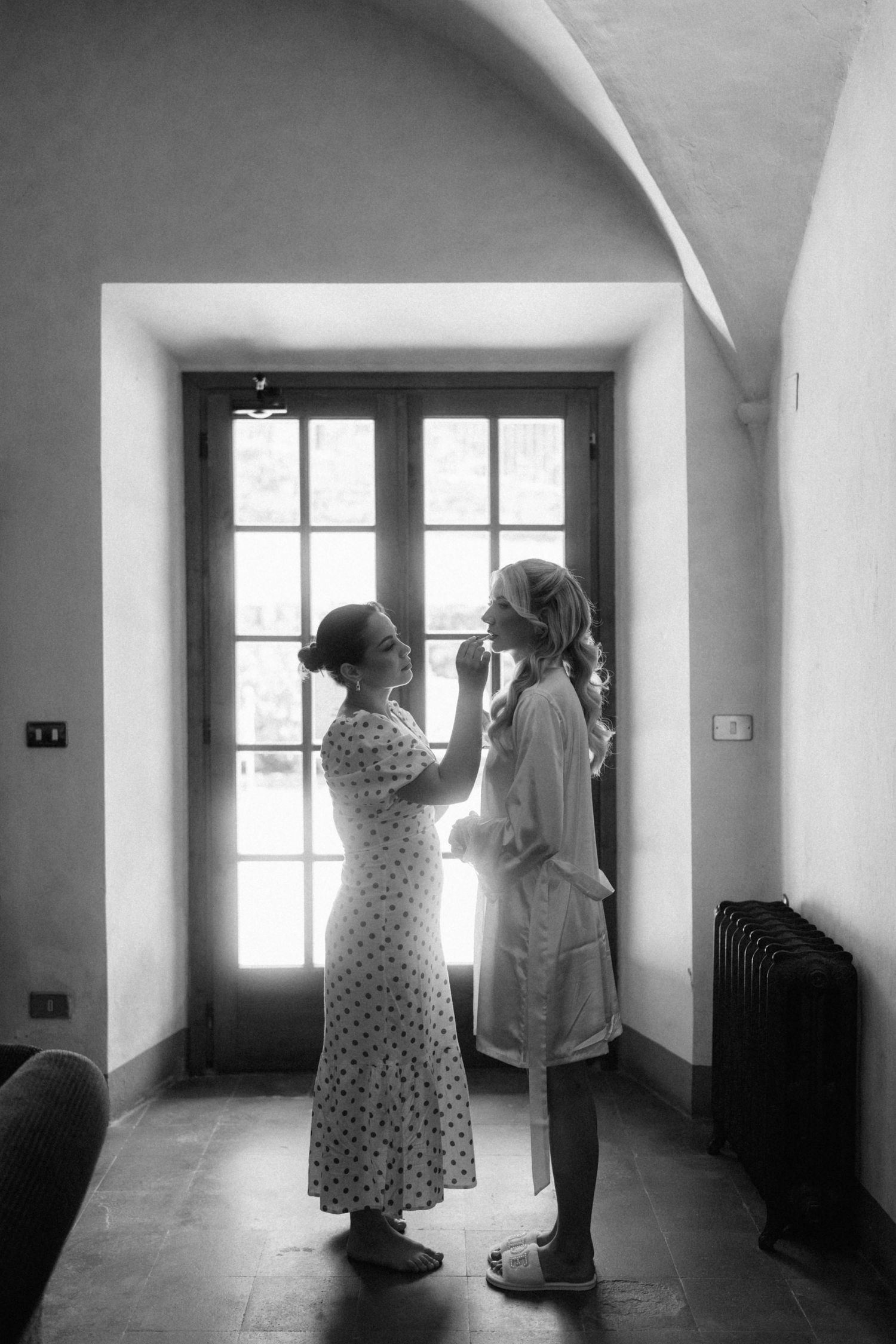 Black and white photo of two women, one helping the other with makeup near a window.