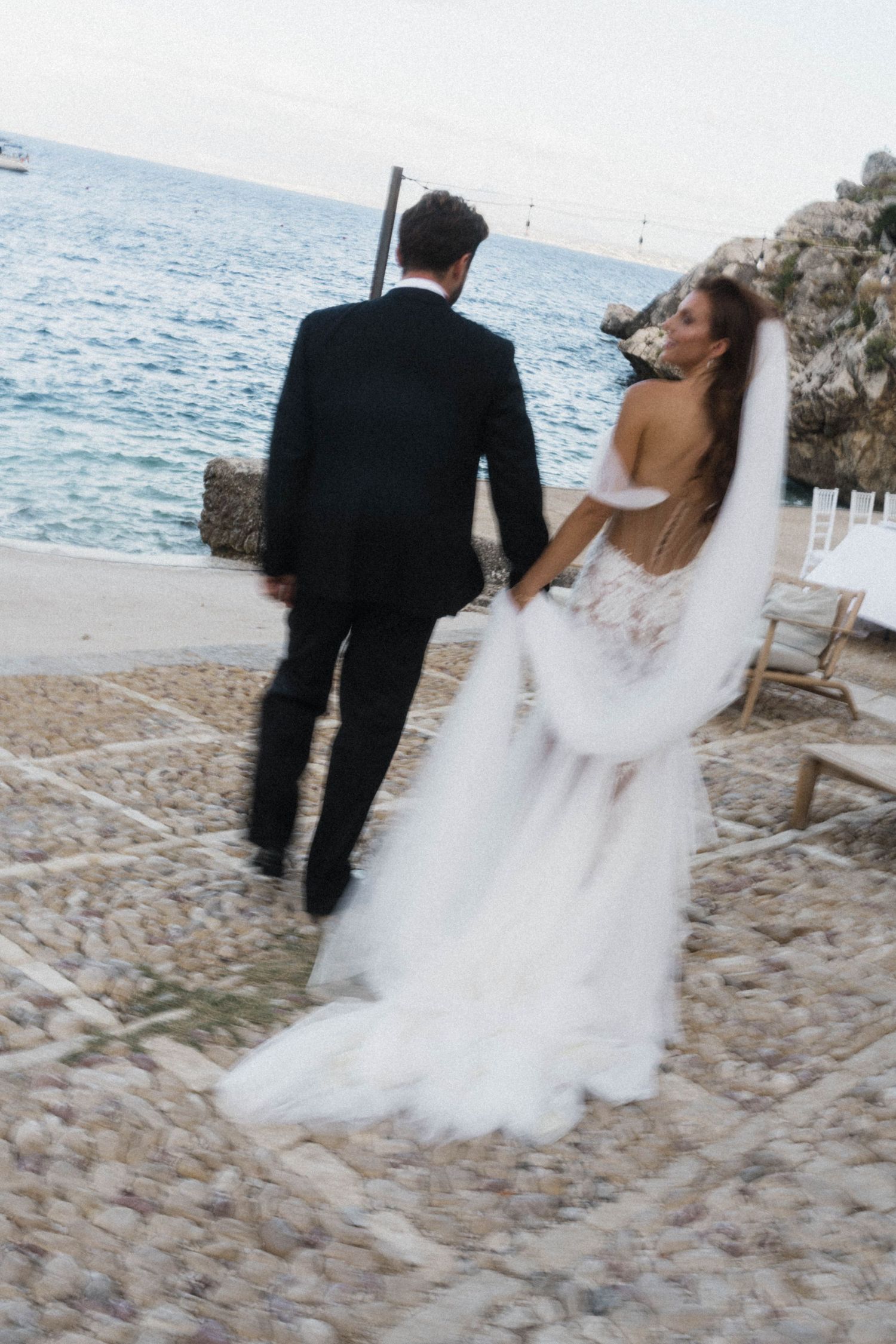 Newlyweds walk hand in hand toward the sea at coastal wedding venue.