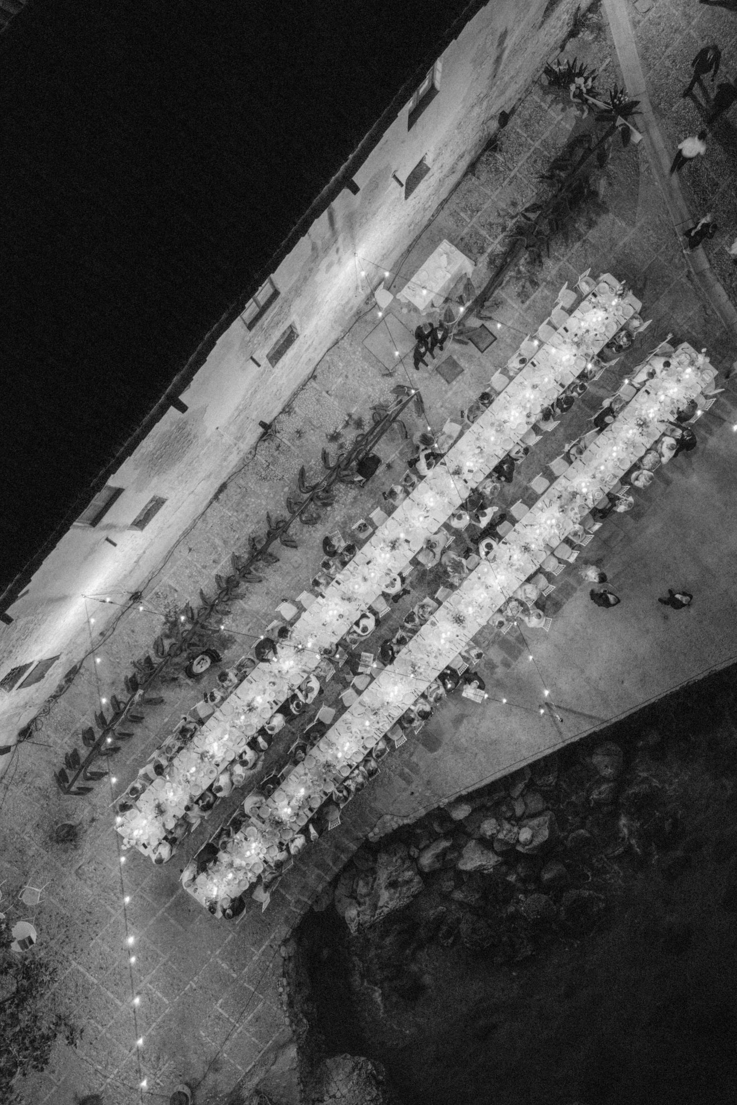 Black and white aerial drone view of long illuminated wedding reception tables arranged outdoors at night.