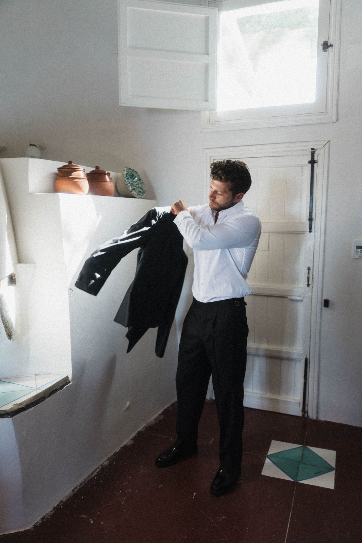 Groom in white shirt holding black suit jacket while getting dressed in rustic whitewashed Mediterranean room.