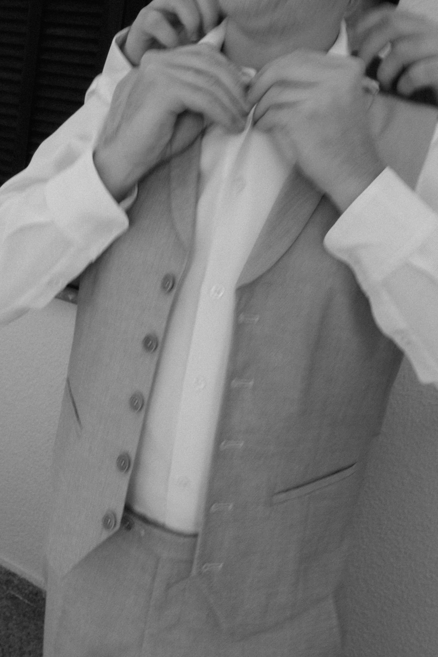 Black and white close-up of groom in vest and white shirt having tie adjusted by helper.