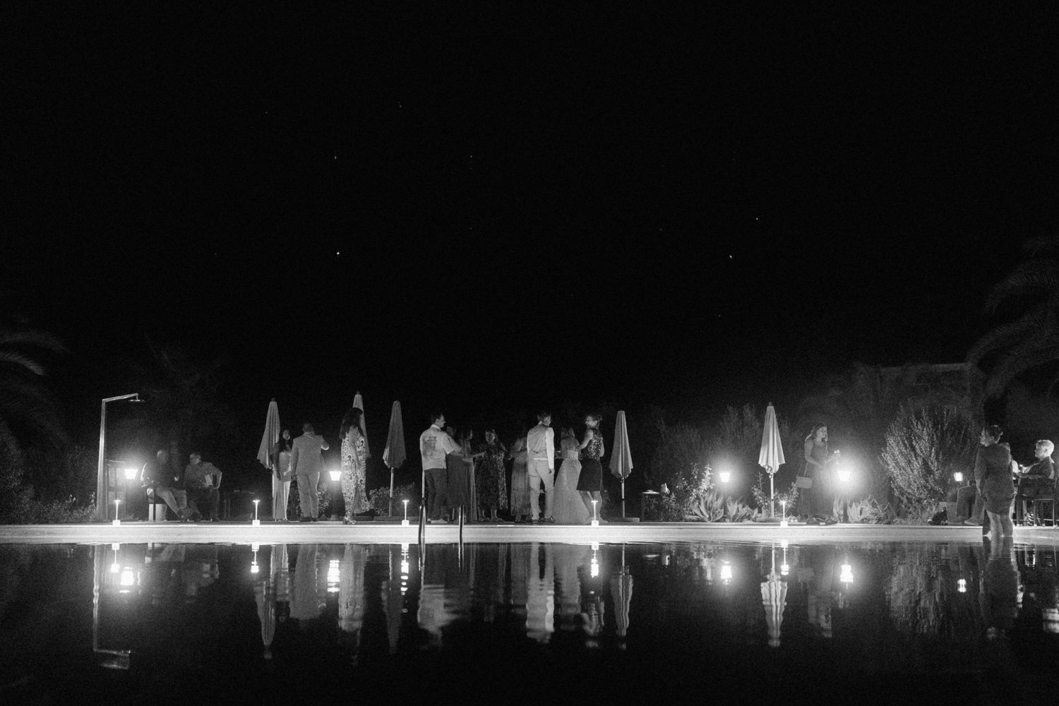 Black and white nighttime photo of wedding guests gathered by a reflective pool surrounded by palm trees.