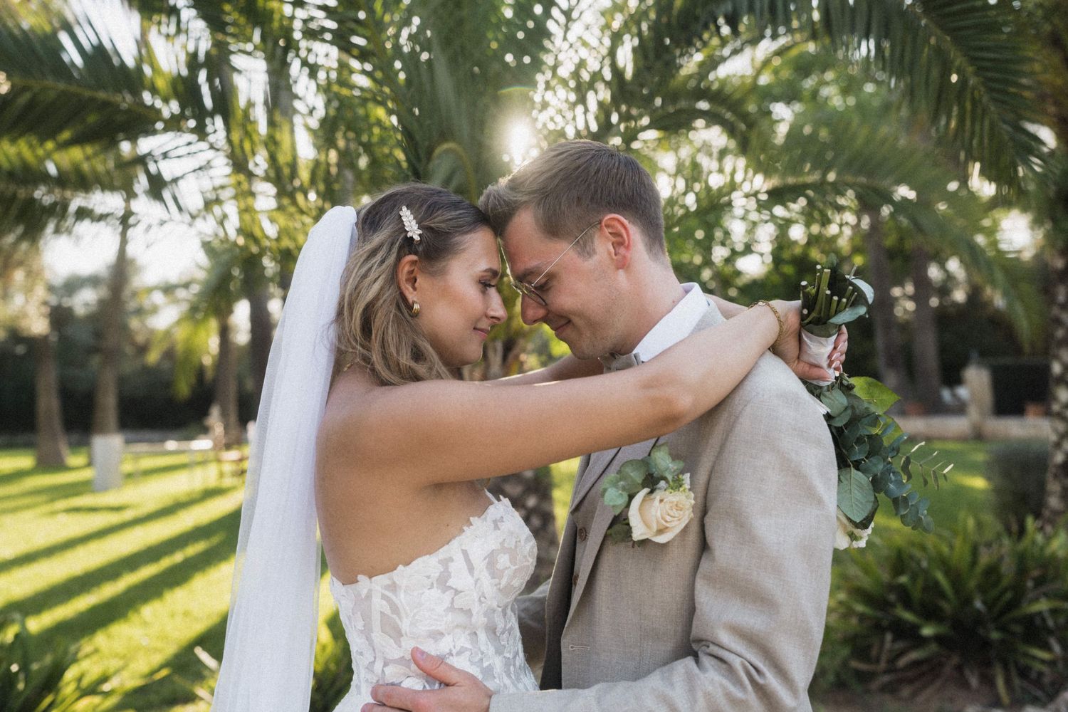 Bride and groom touching foreheads romantically under golden sunlight surrounded by lush palm trees.