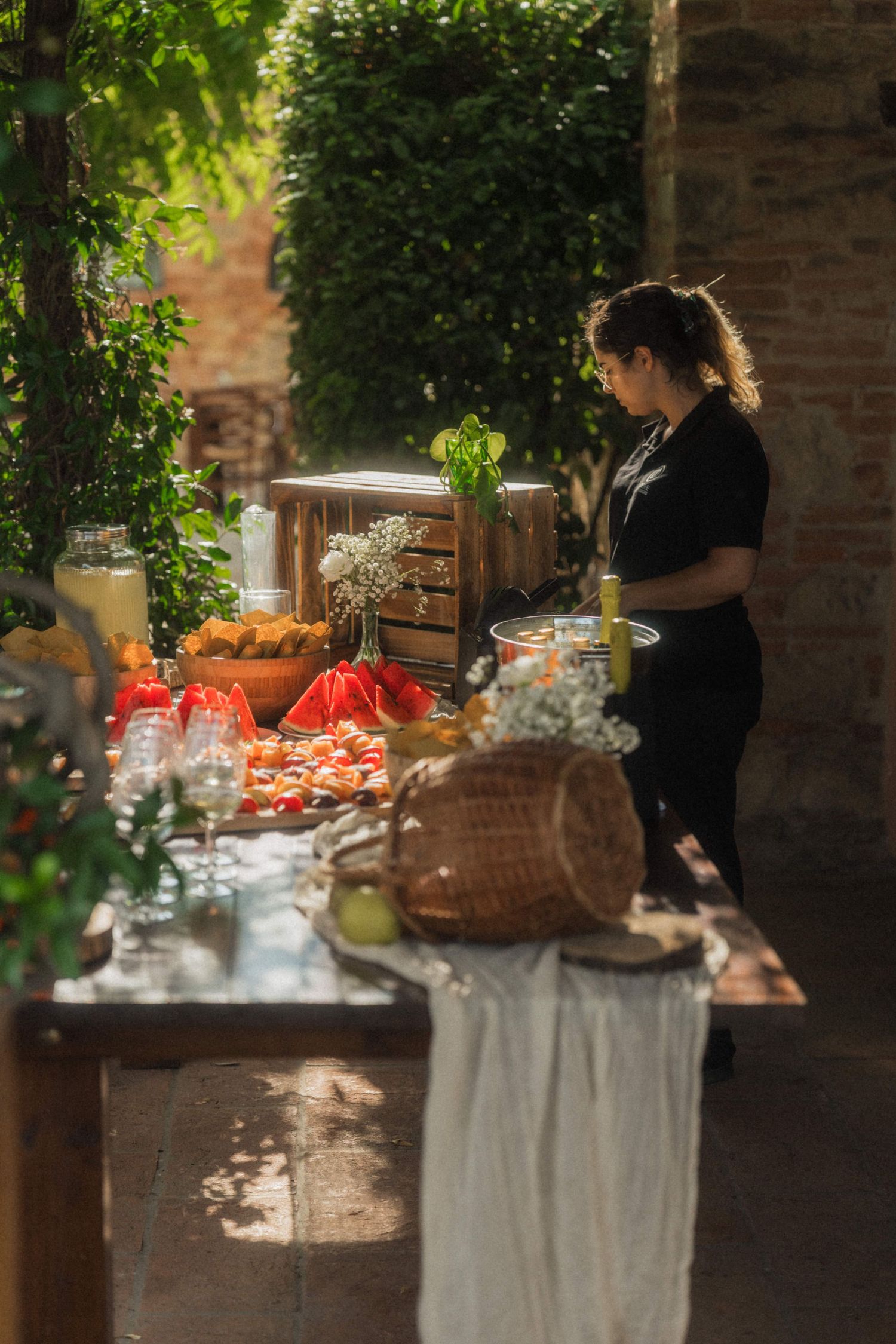 Catering staff arranges colorful food spread on outdoor buffet table at sunny garden wedding reception.