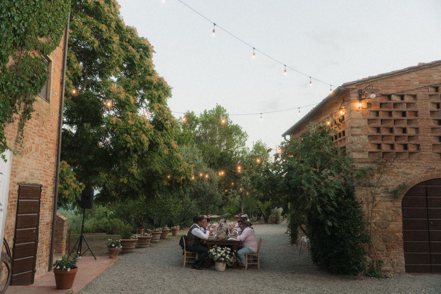 Intimate outdoor wedding dinner at dusk with string lights between rustic stone buildings and ivy-covered walls.