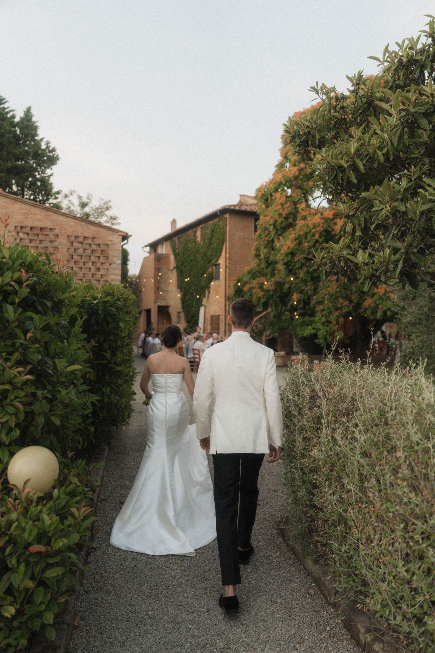 Bride and groom walking hand in hand along a garden path at a Tuscan villa wedding venue at dusk.