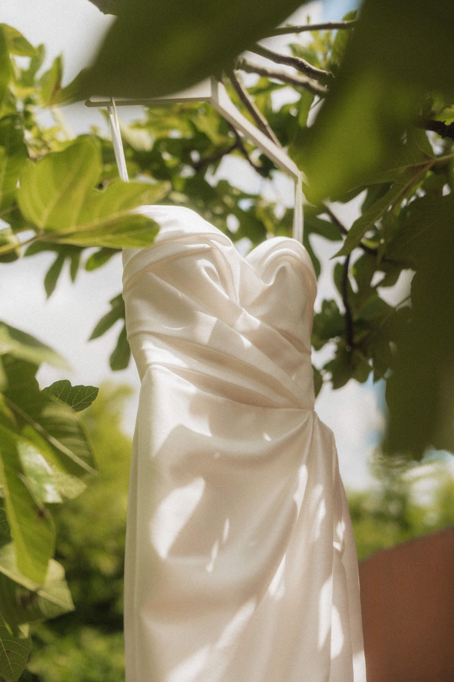 Elegant white satin strapless wedding dress hanging on a hanger among lush green tree branches outdoors.