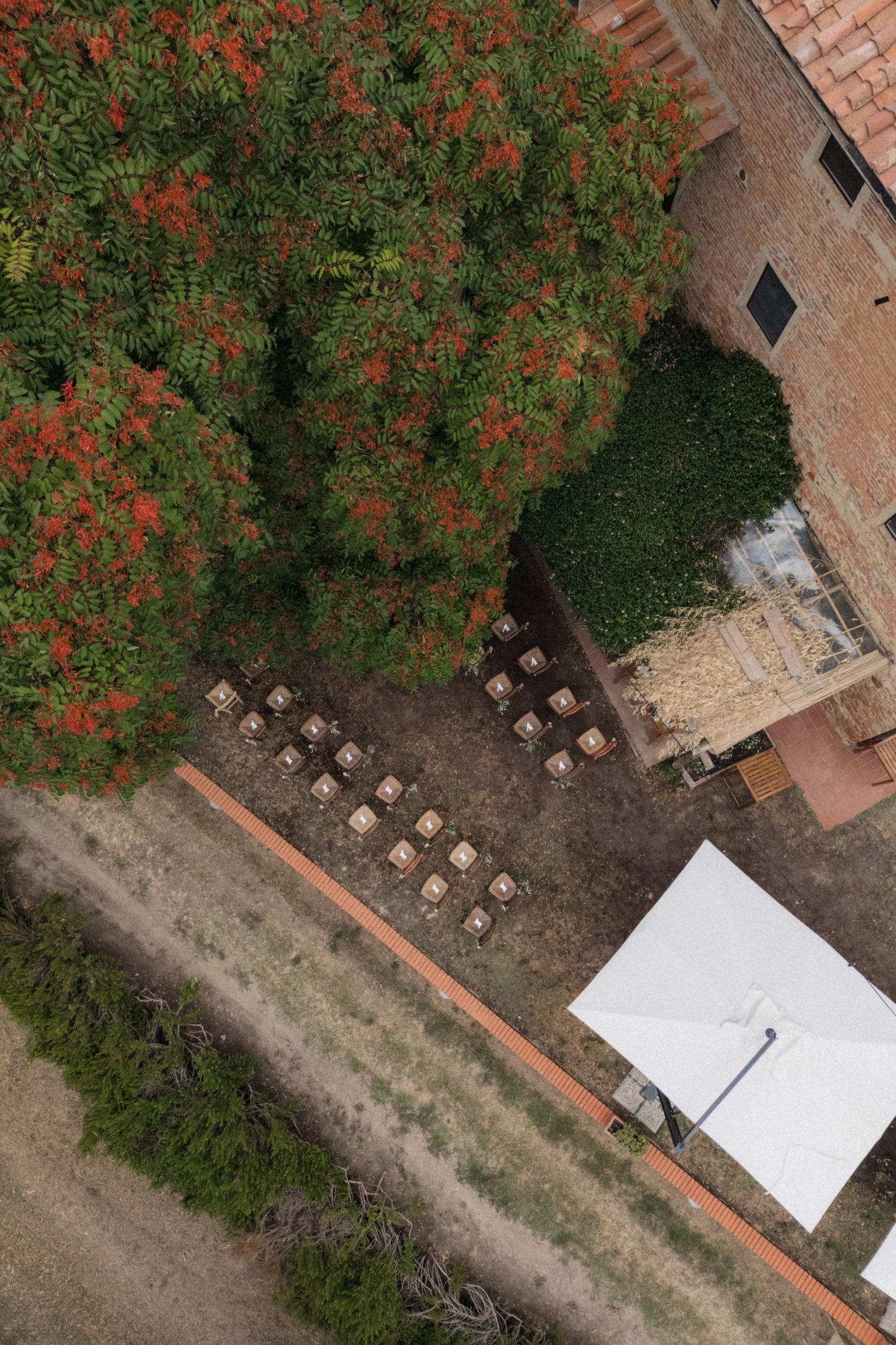 Aerial view of outdoor wedding ceremony chairs arranged near blooming red tree at rustic Tuscan estate.