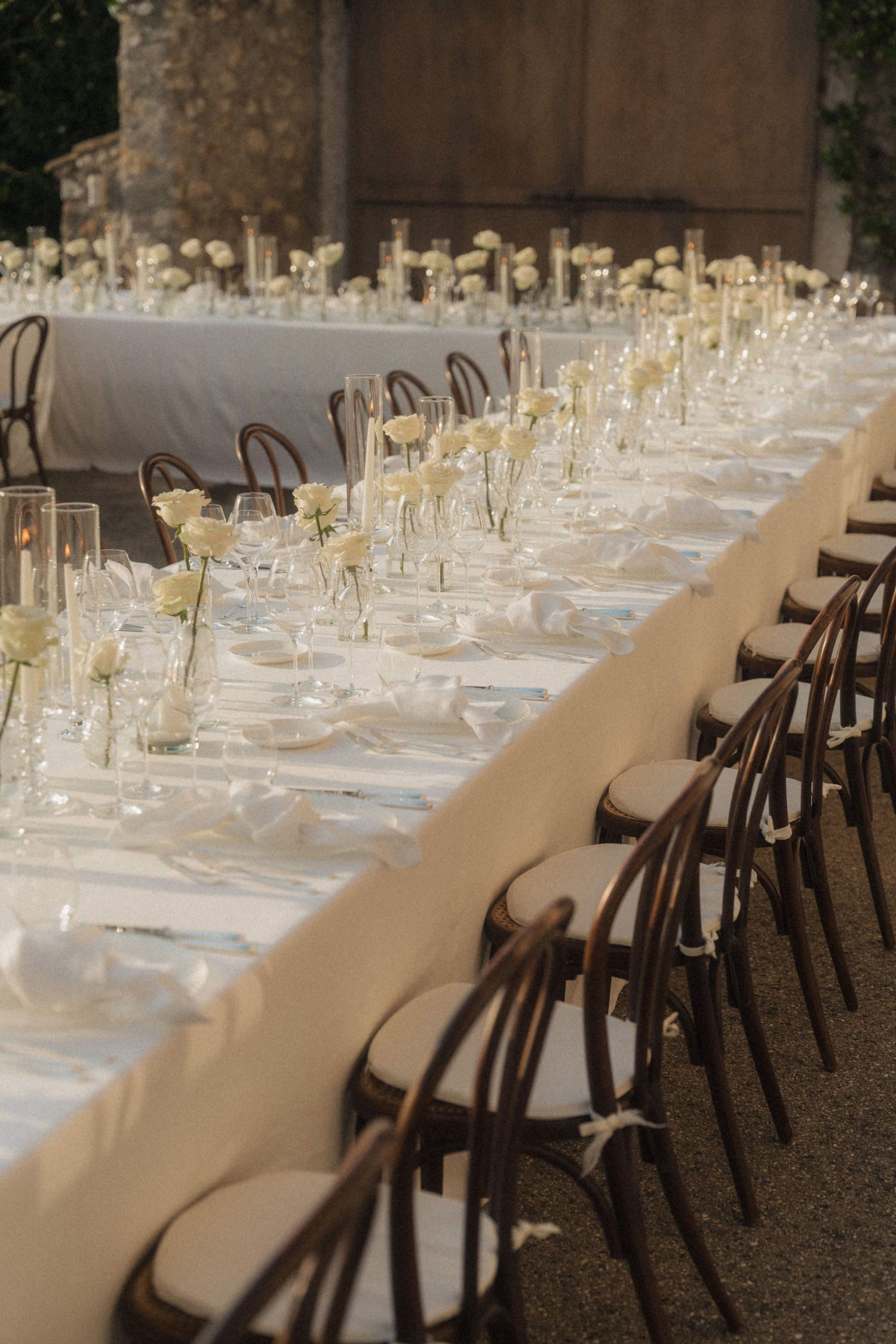 Elegant long wedding reception table with white roses, candles, and dark bentwood chairs.