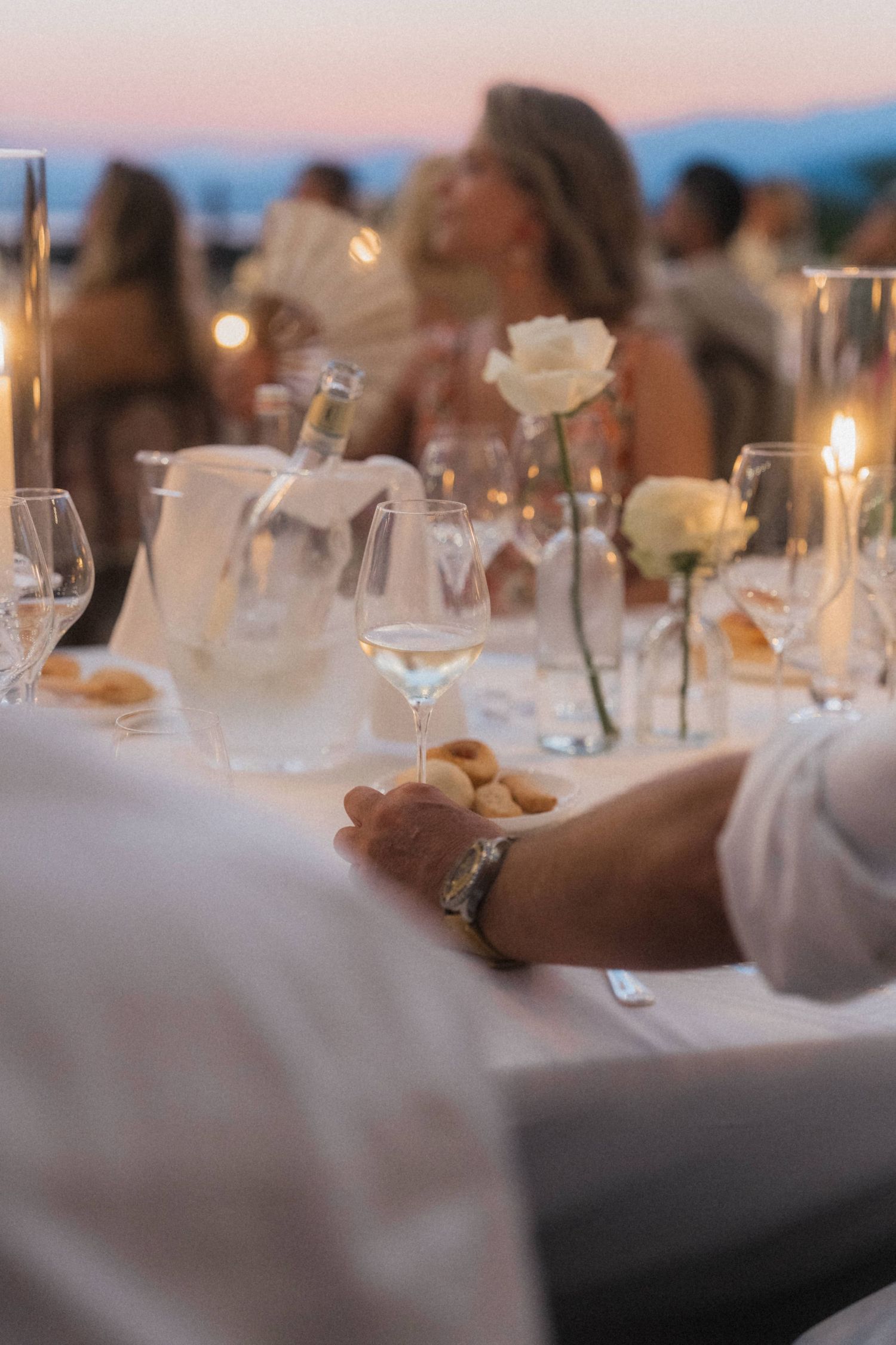 Wedding reception dinner table at dusk with white roses, candles, and wine glasses.