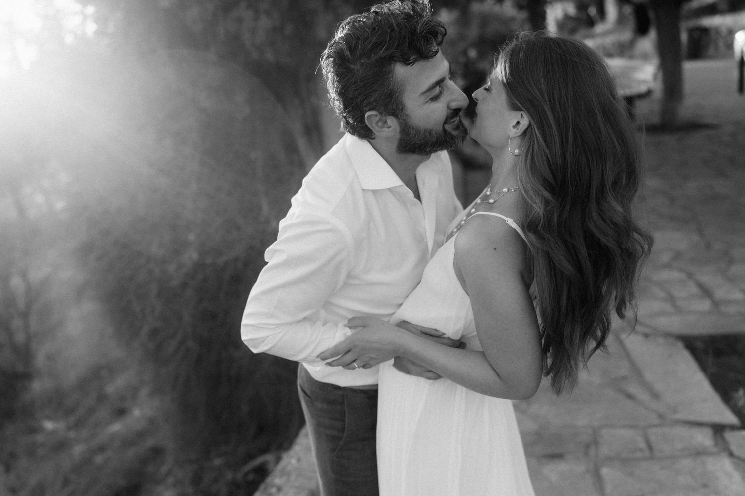 Black and white photo of a romantic couple sharing a tender kiss outdoors in a garden setting.