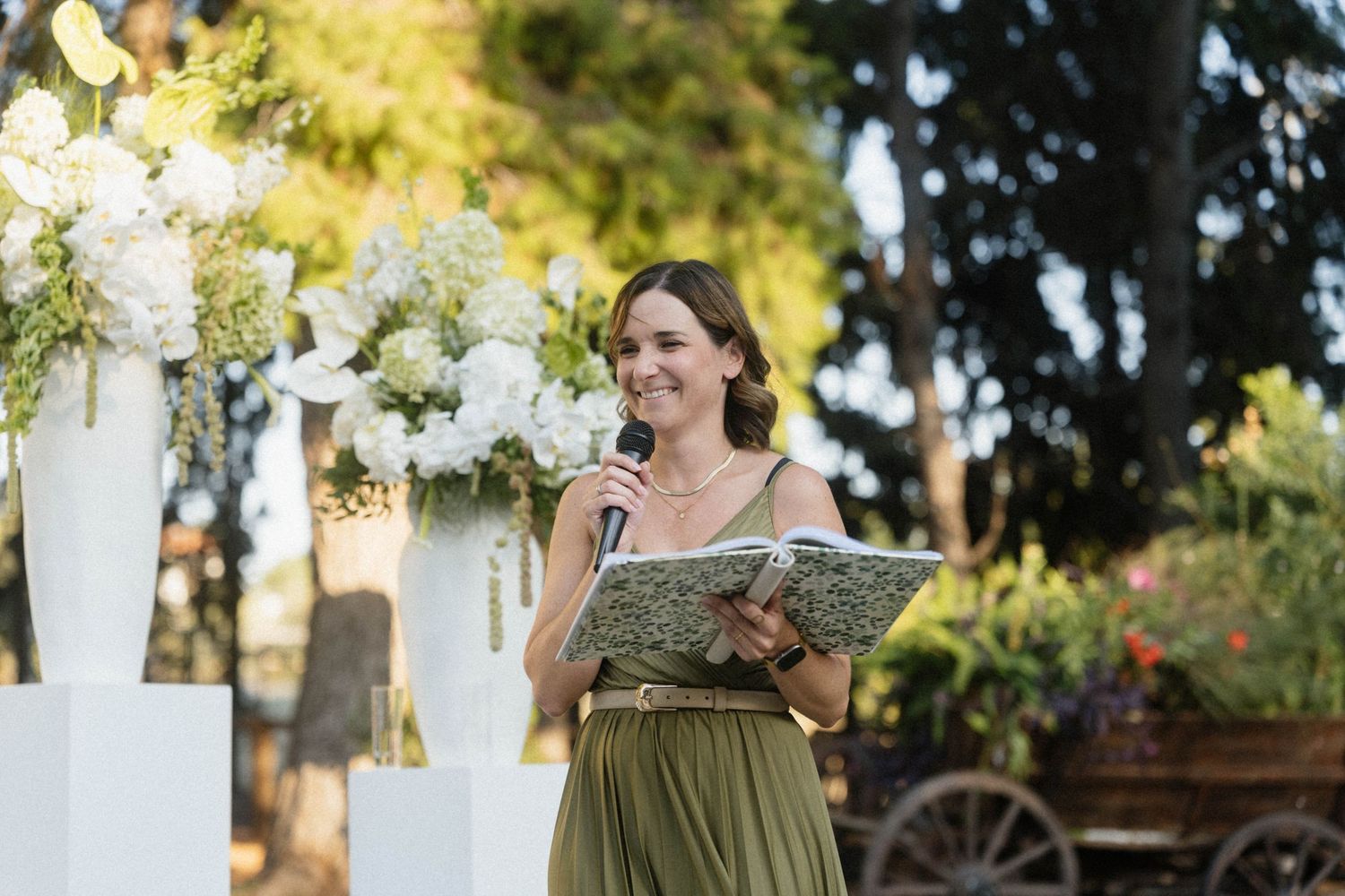 Woman in green dress holding microphone and book speaks at outdoor wedding ceremony with white floral arrangements.
