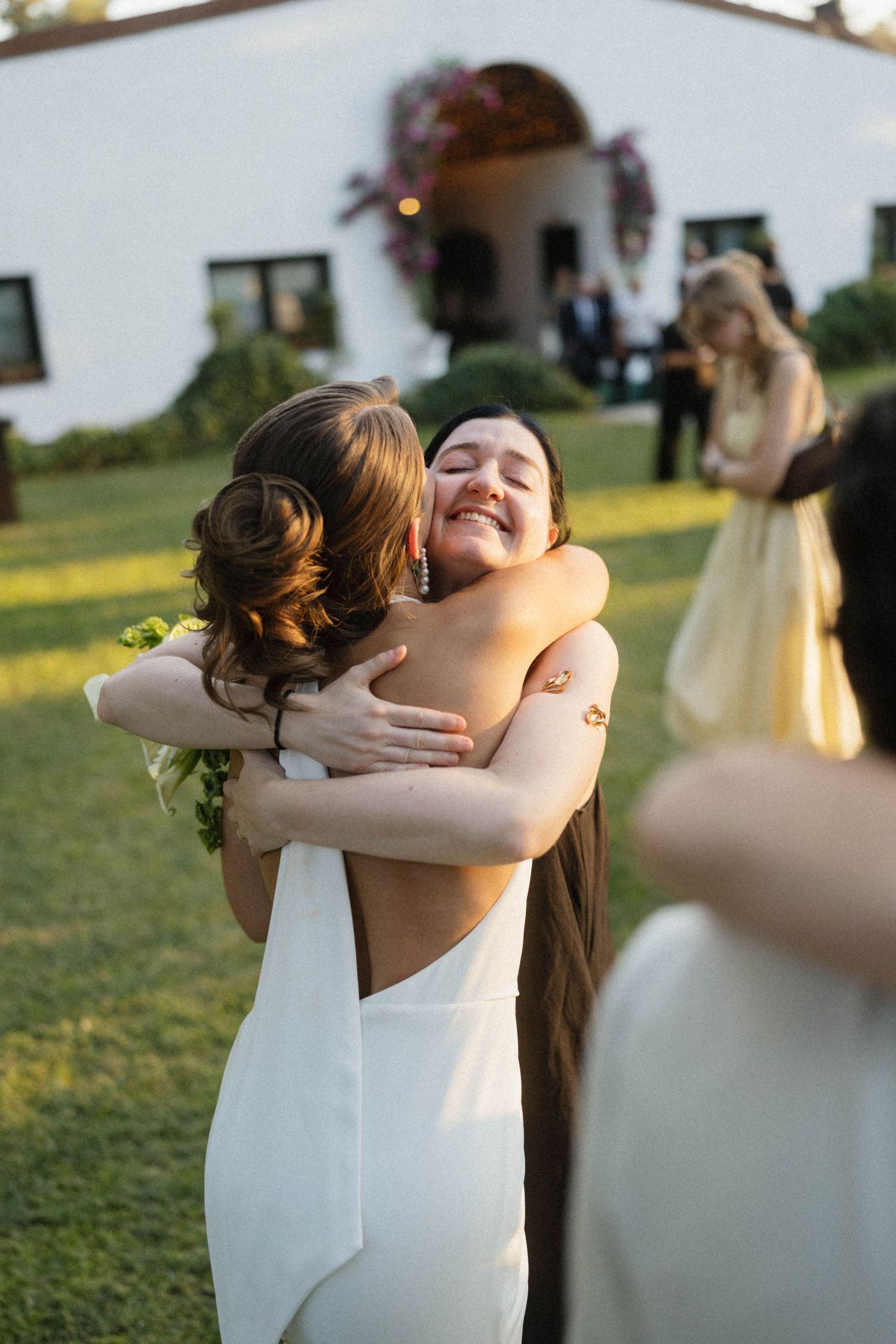 Bride in white gown shares joyful hug with smiling guest outdoors at elegant wedding venue.