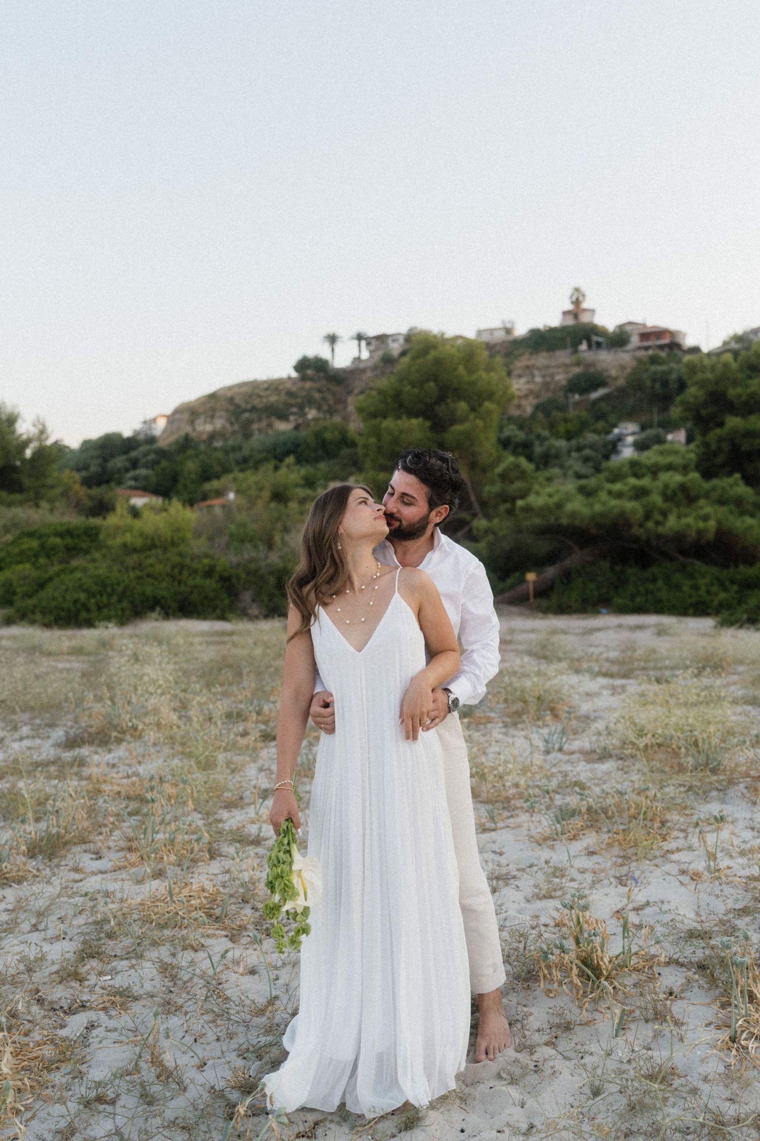 Couple in white wedding attire embrace on sandy beach with lush green hillside backdrop at dusk.