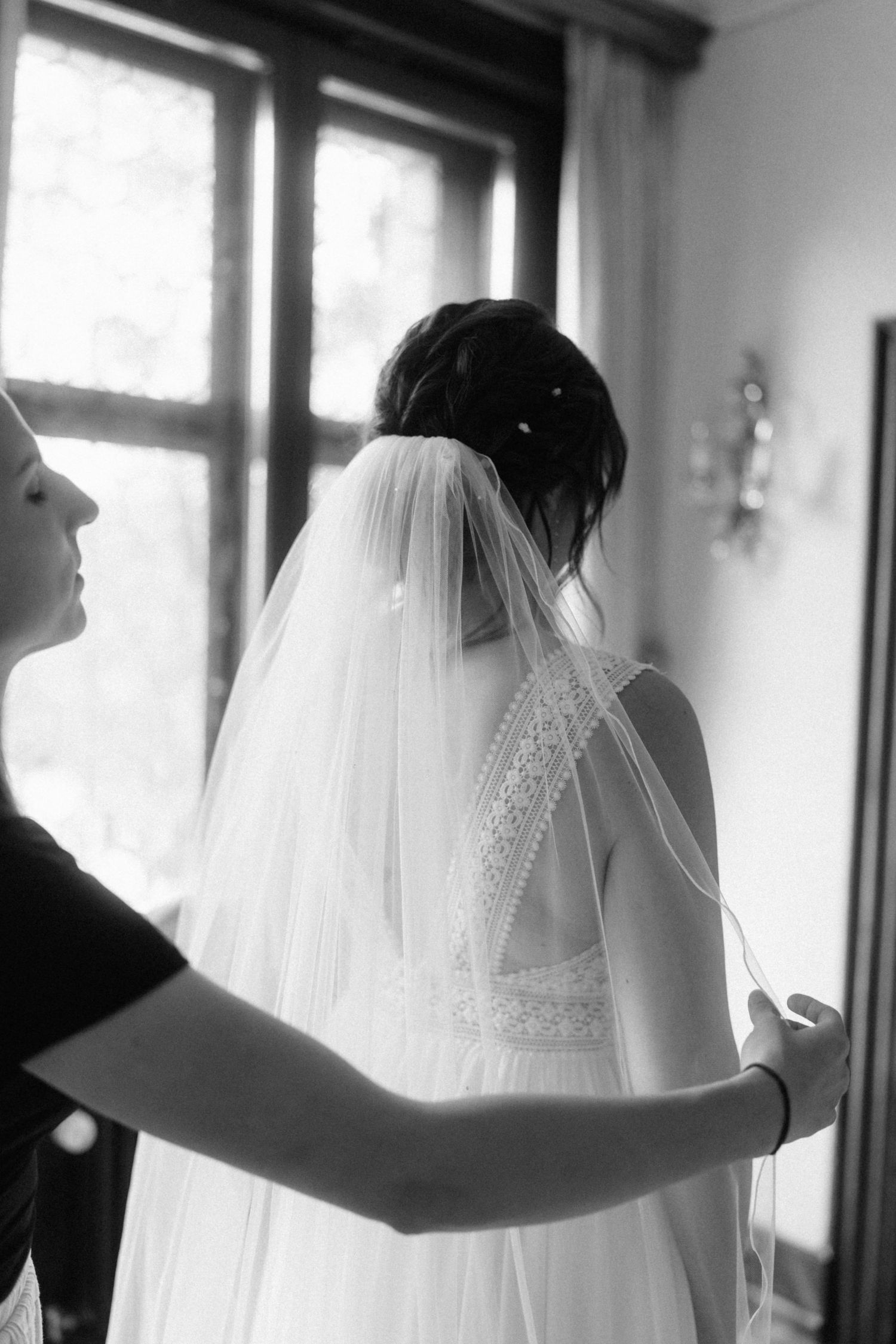 Black and white photo of bridesmaid adjusting wedding veil on bride in lace back gown by window.