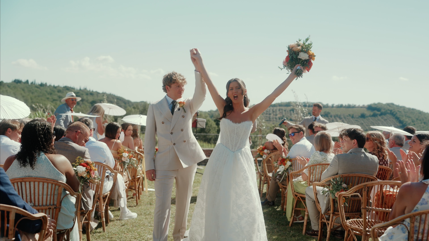 Joyful newlyweds celebrate at outdoor wedding ceremony, bride in white gown holding bouquet, groom in tan suit.