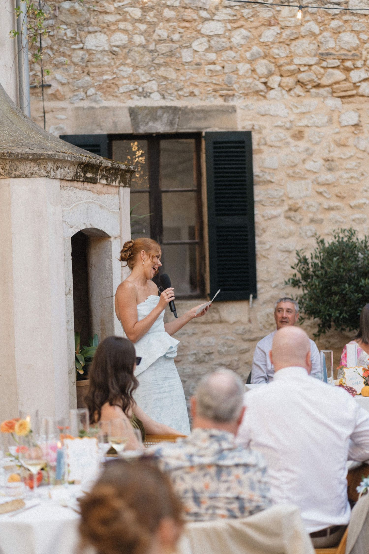 Bride in white dress giving speech at outdoor wedding reception beside rustic stone building.